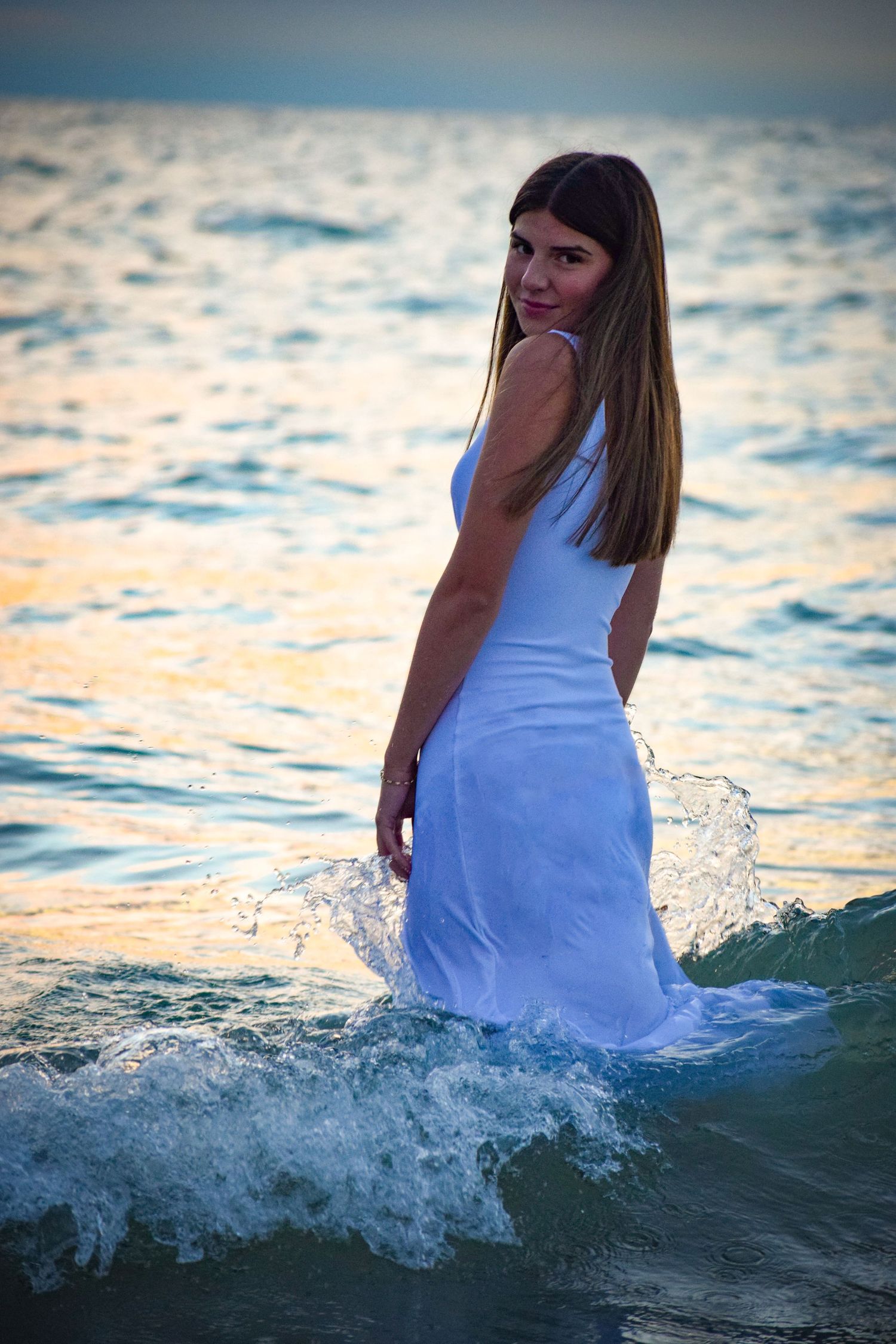 Person in white dress sitting in ocean waves during sunset.