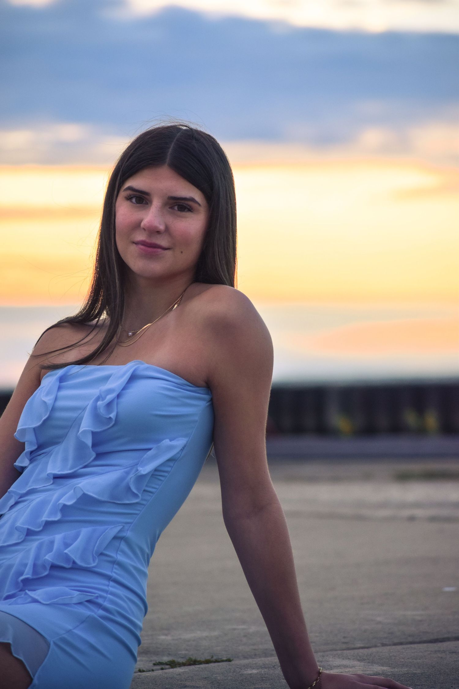 Woman in light blue ruffled dress poses against sunset sky.
