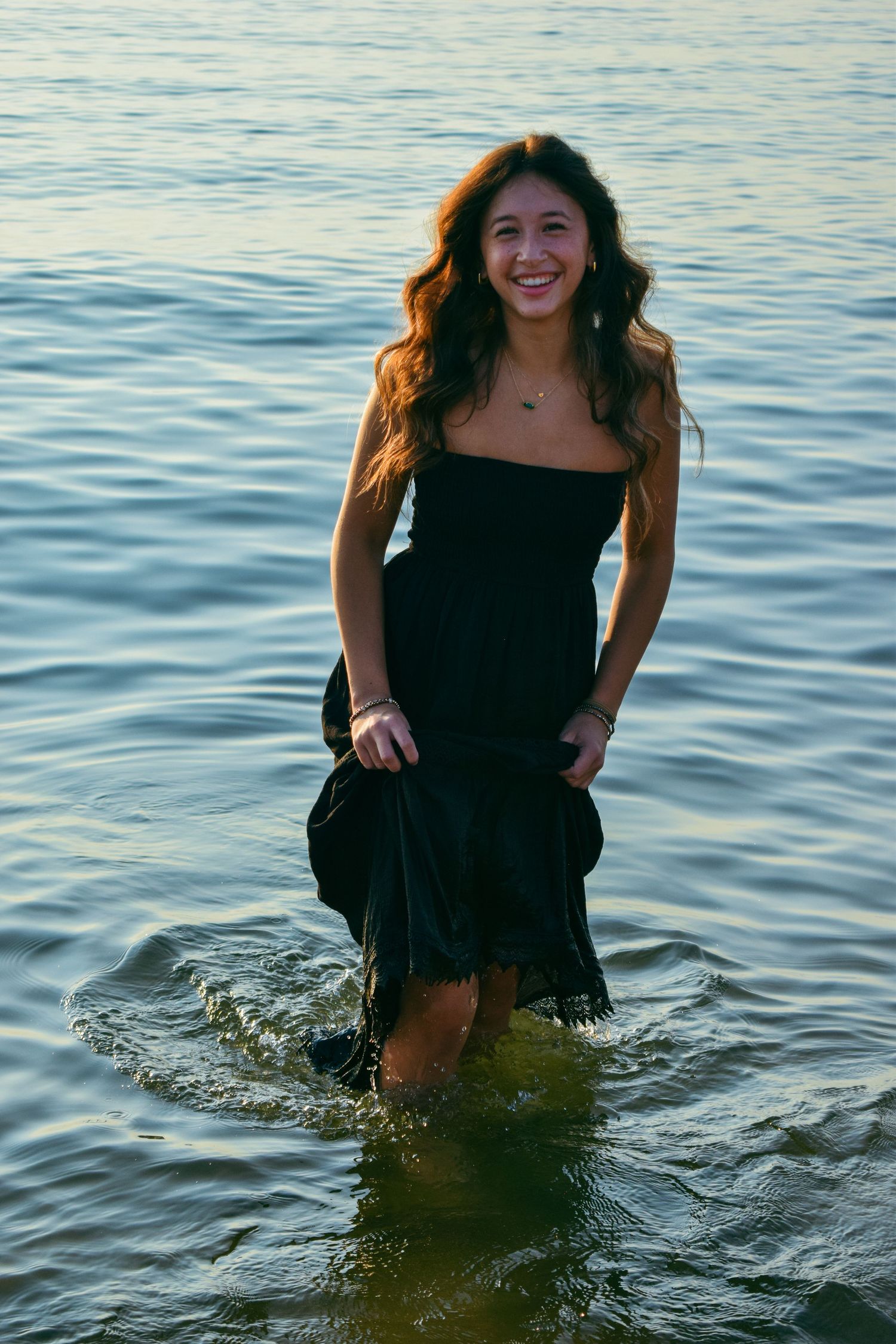Woman in black dress plays in ocean water during beach photoshoot.