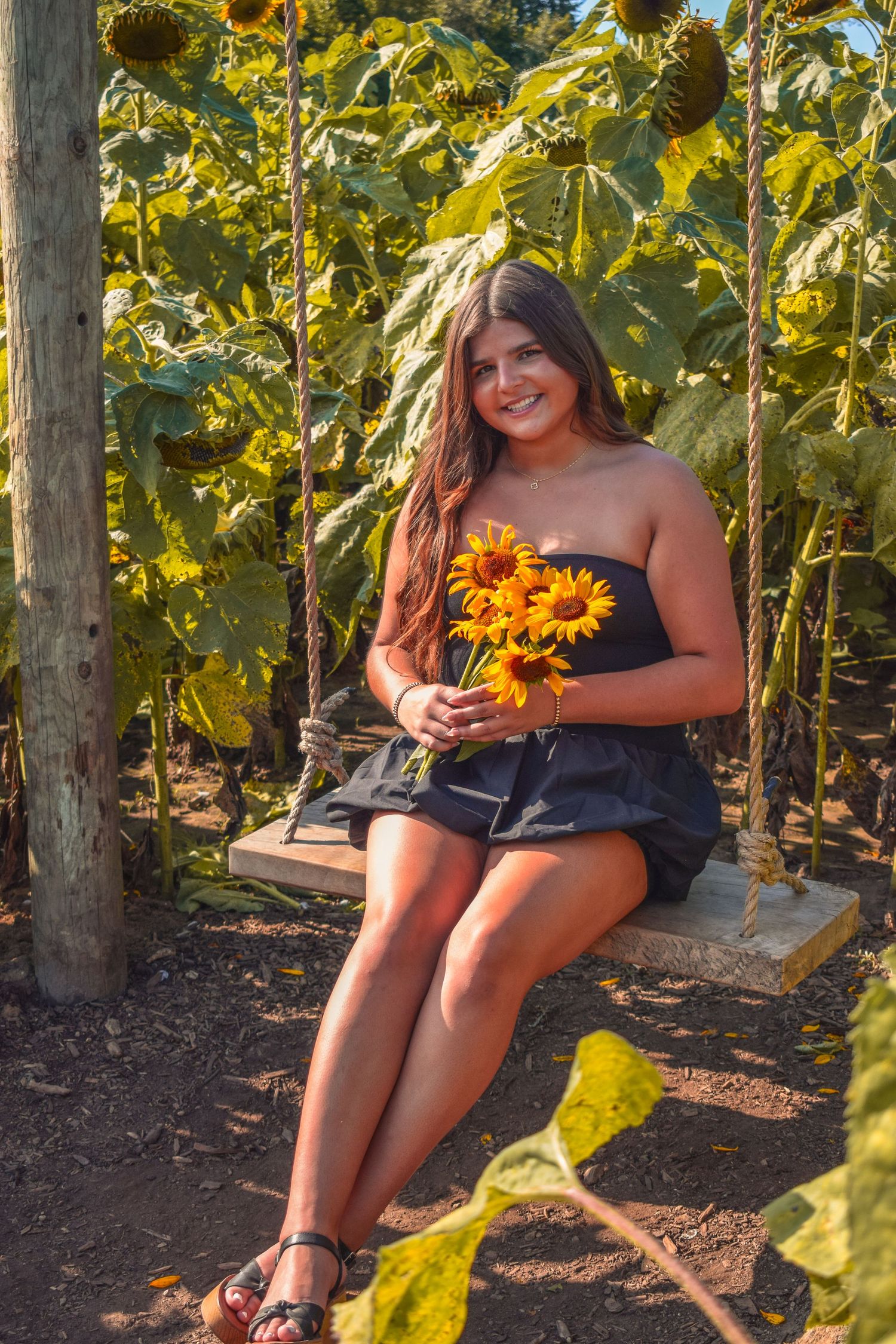 Woman in black top sits on swing surrounded by tall sunflowers in garden setting.