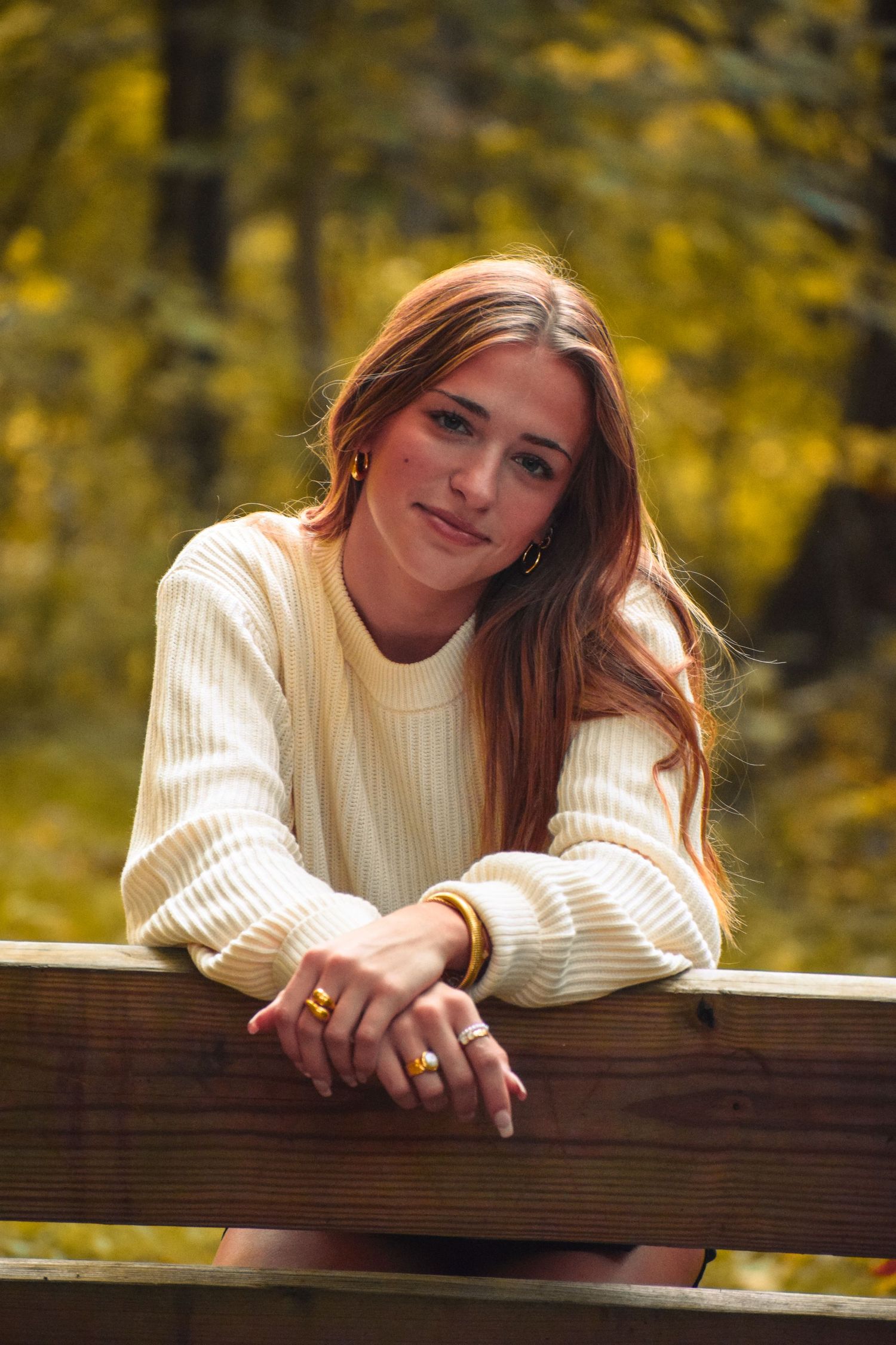 Person in white sweater leans on wooden railing with autumn foliage behind.