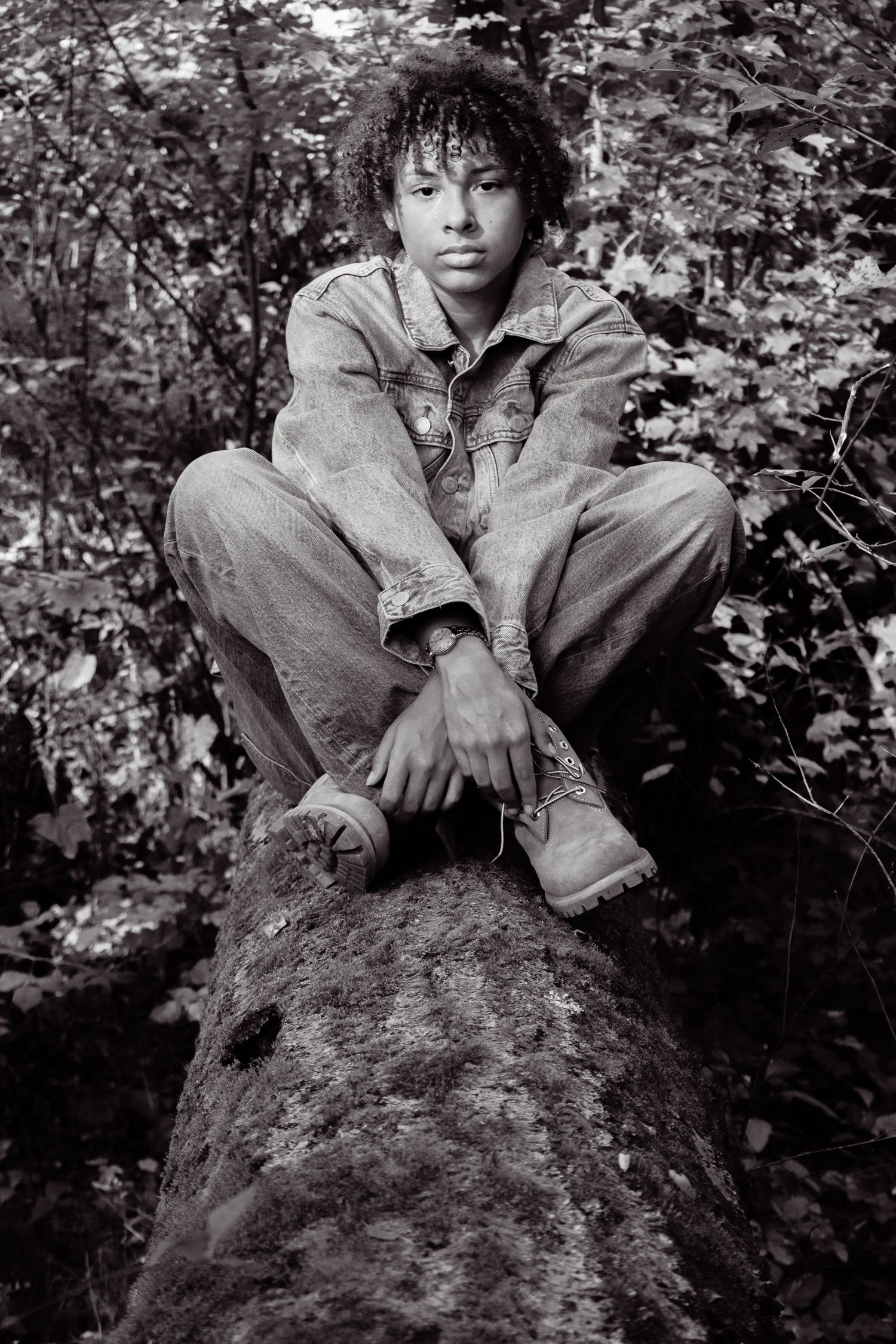 Figure in denim sits casually on mossy rock in black and white forest setting.
