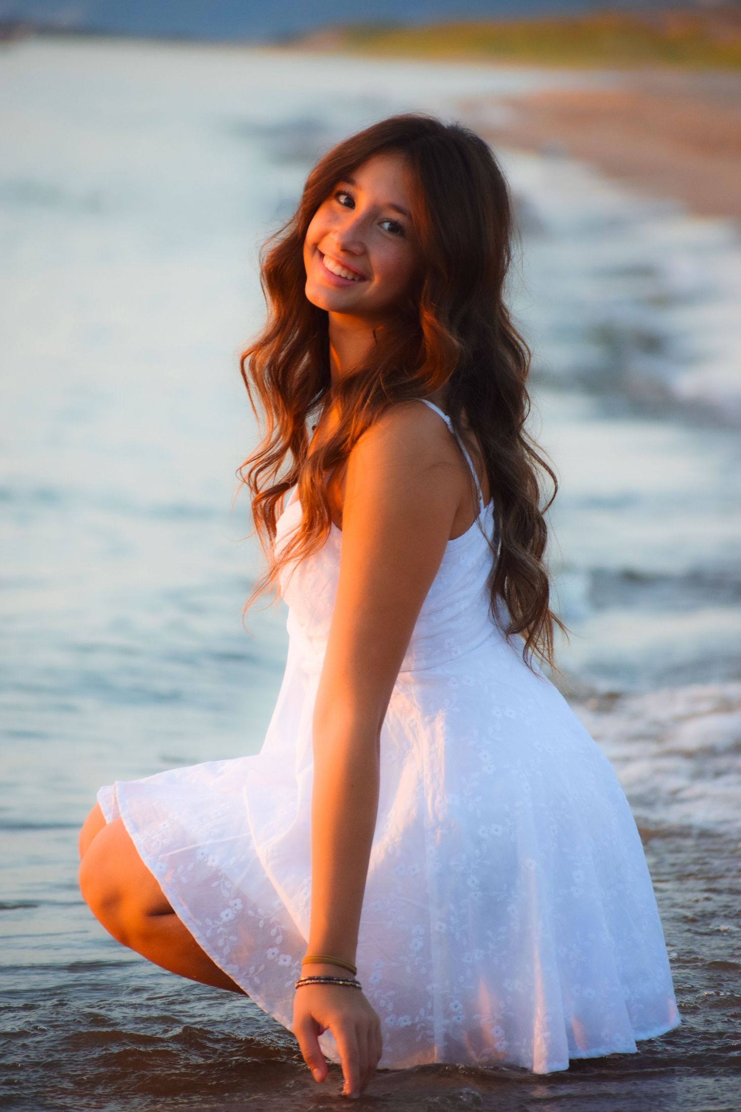 A person in a flowing white dress poses gracefully on a beach at sunset creating a dreamy coastal scene.