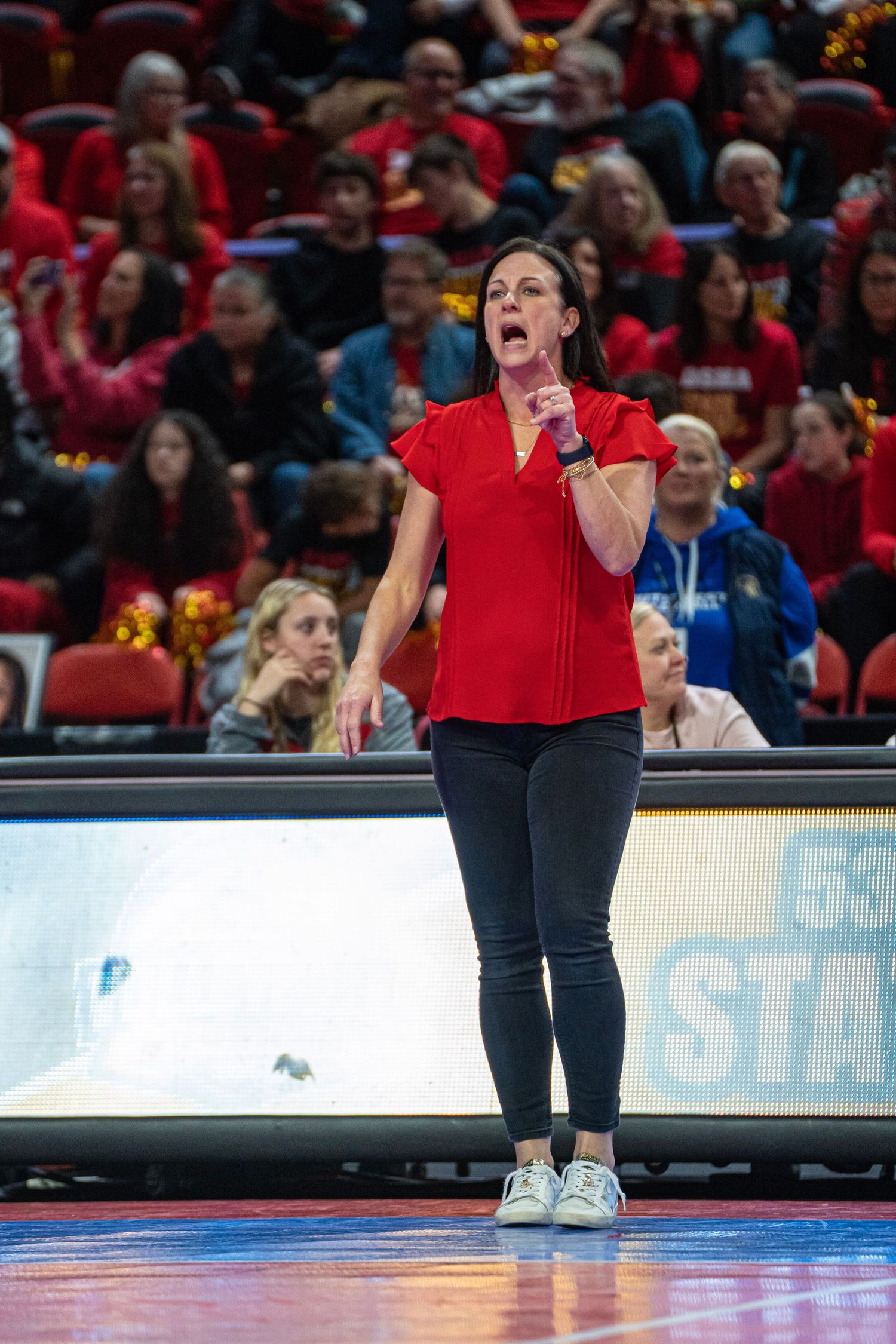 A performer in red attire kneels on a basketball court while singing during a live performance.