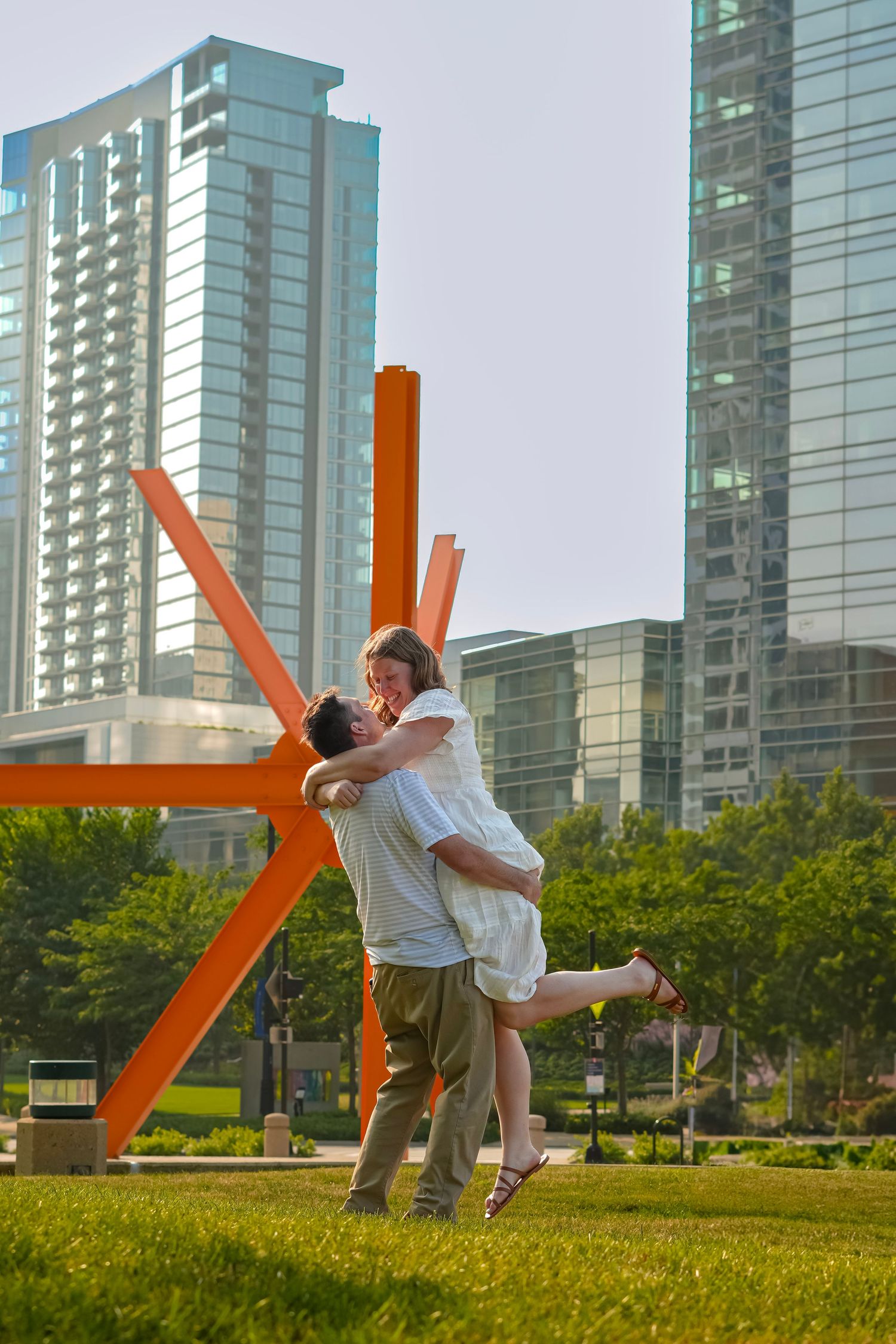 Playful dance moves performed on a lawn with orange sculptures and skyscrapers in the cityscape.