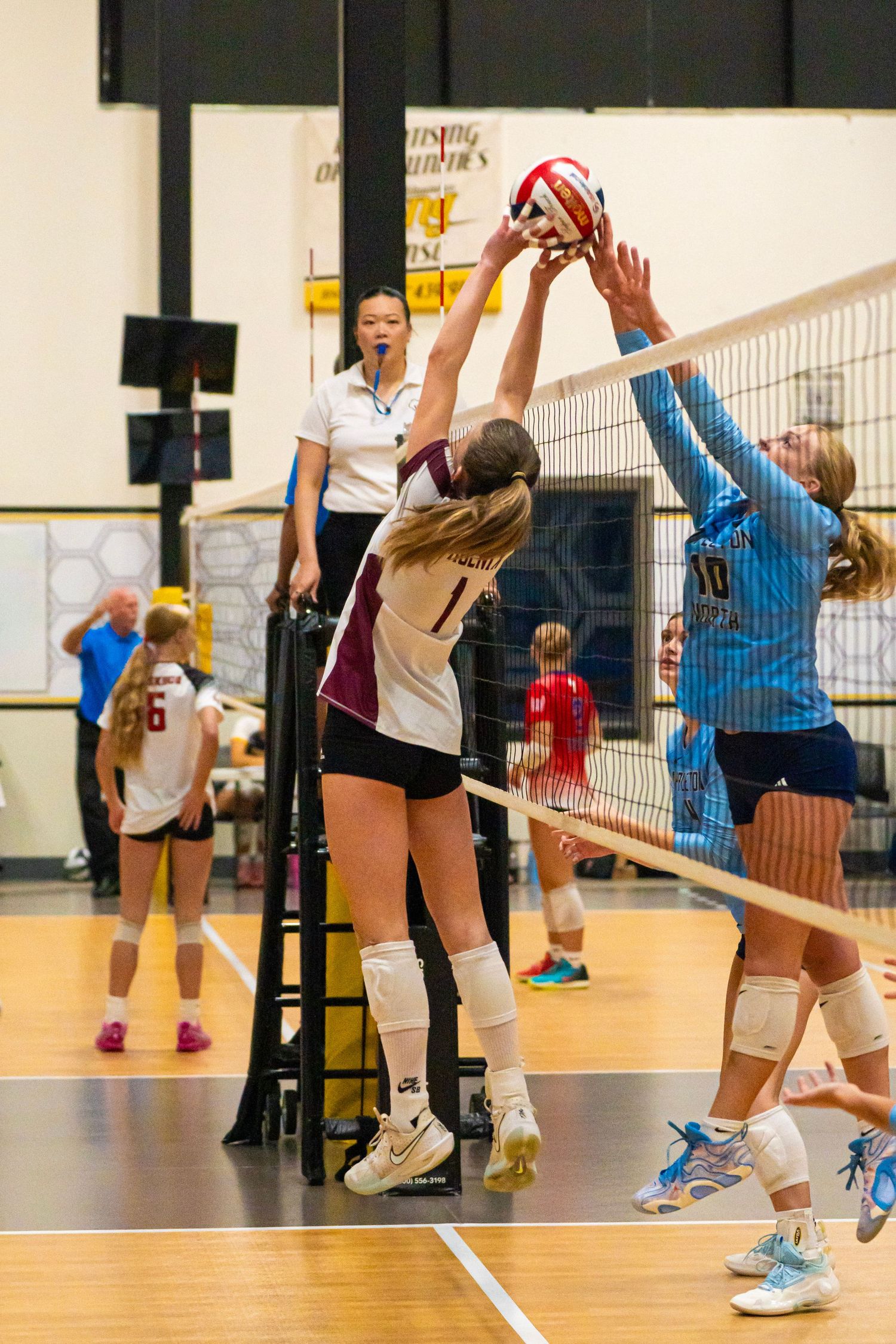 Youth volleyball players jump at net during competitive game action.