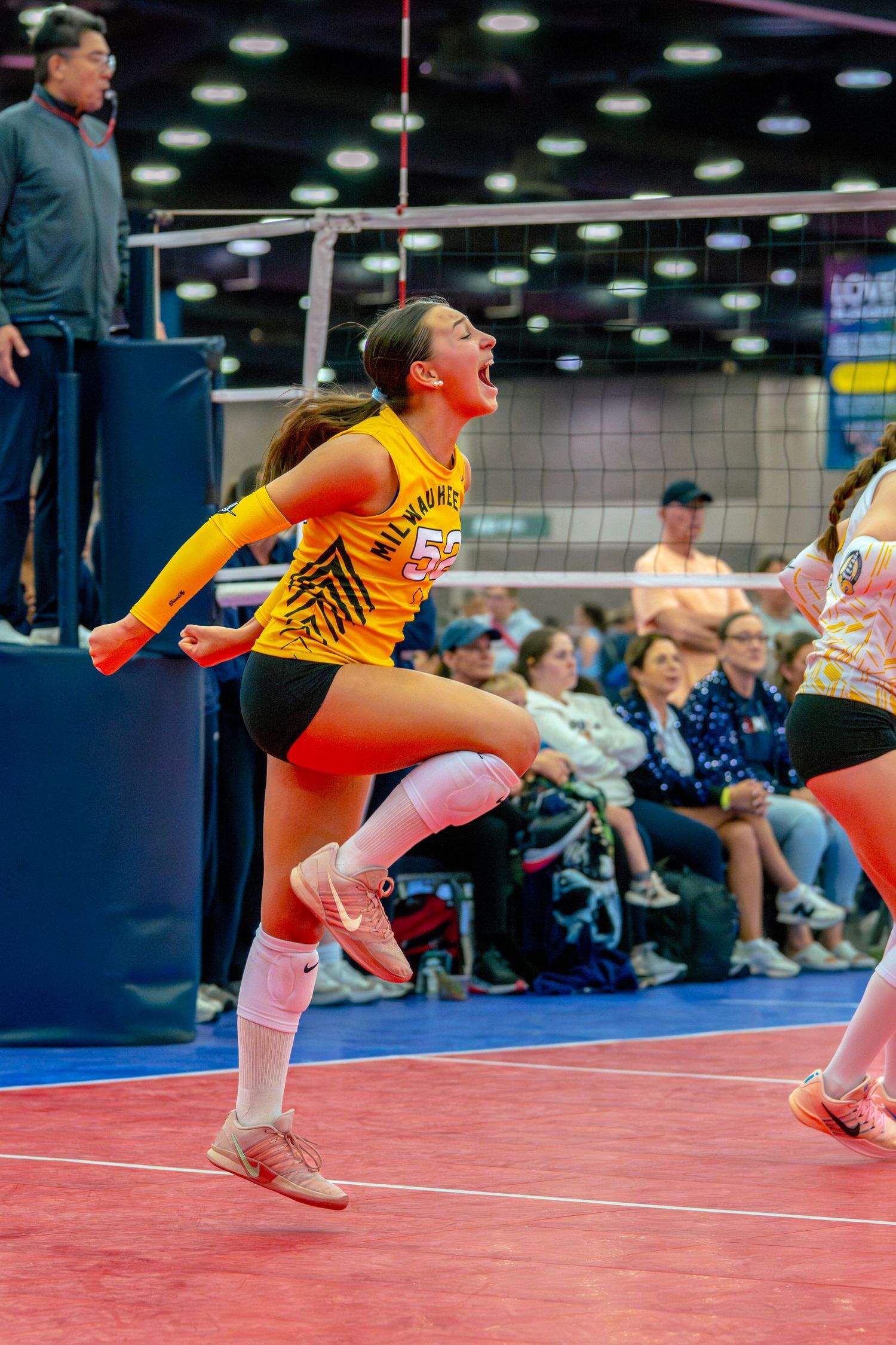 Young female volleyball player in yellow jersey celebrates mid-game at indoor tournament, crowd visible in background.
