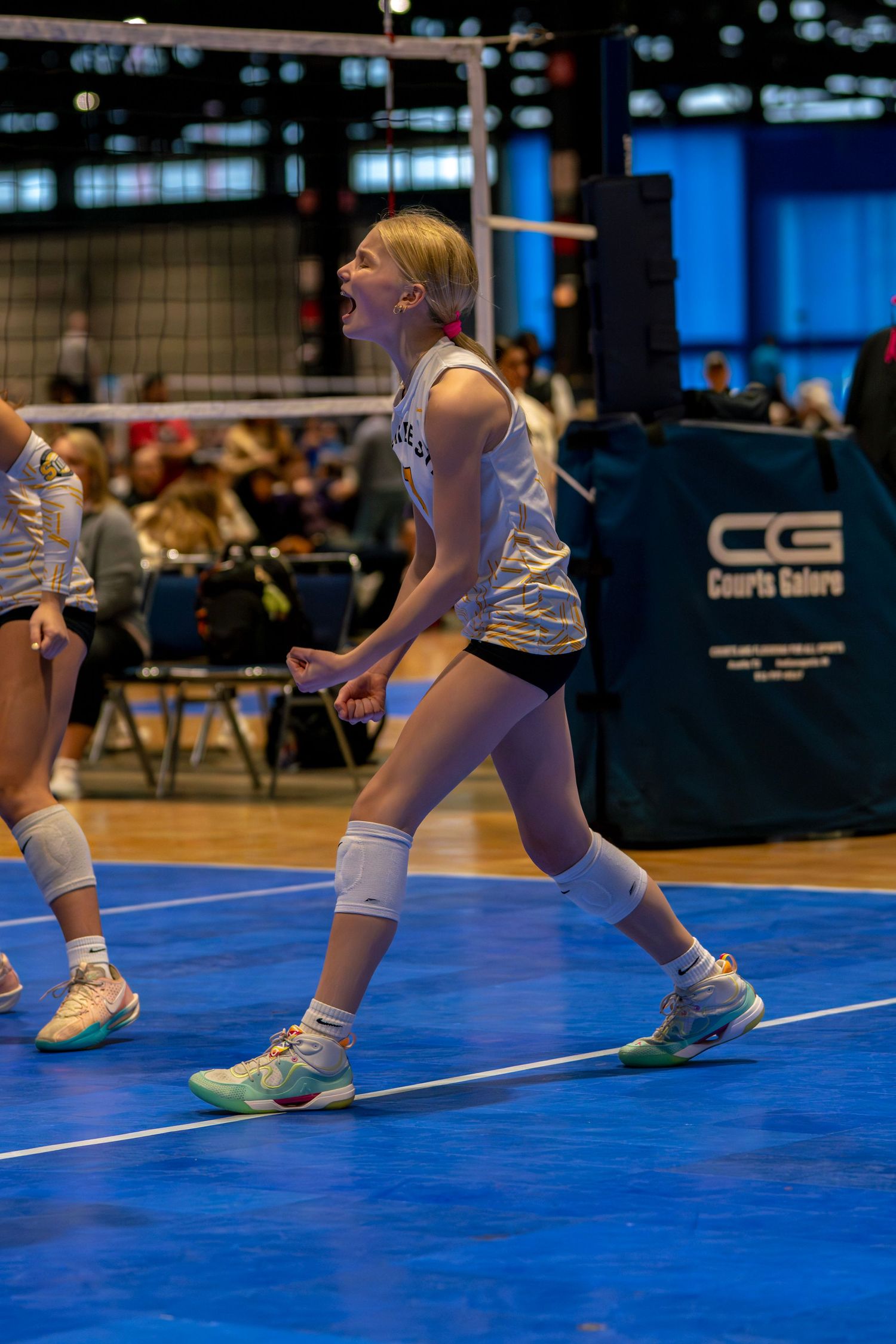 Young female volleyball player celebrates on blue court during indoor tournament, wearing white jersey and black shorts.