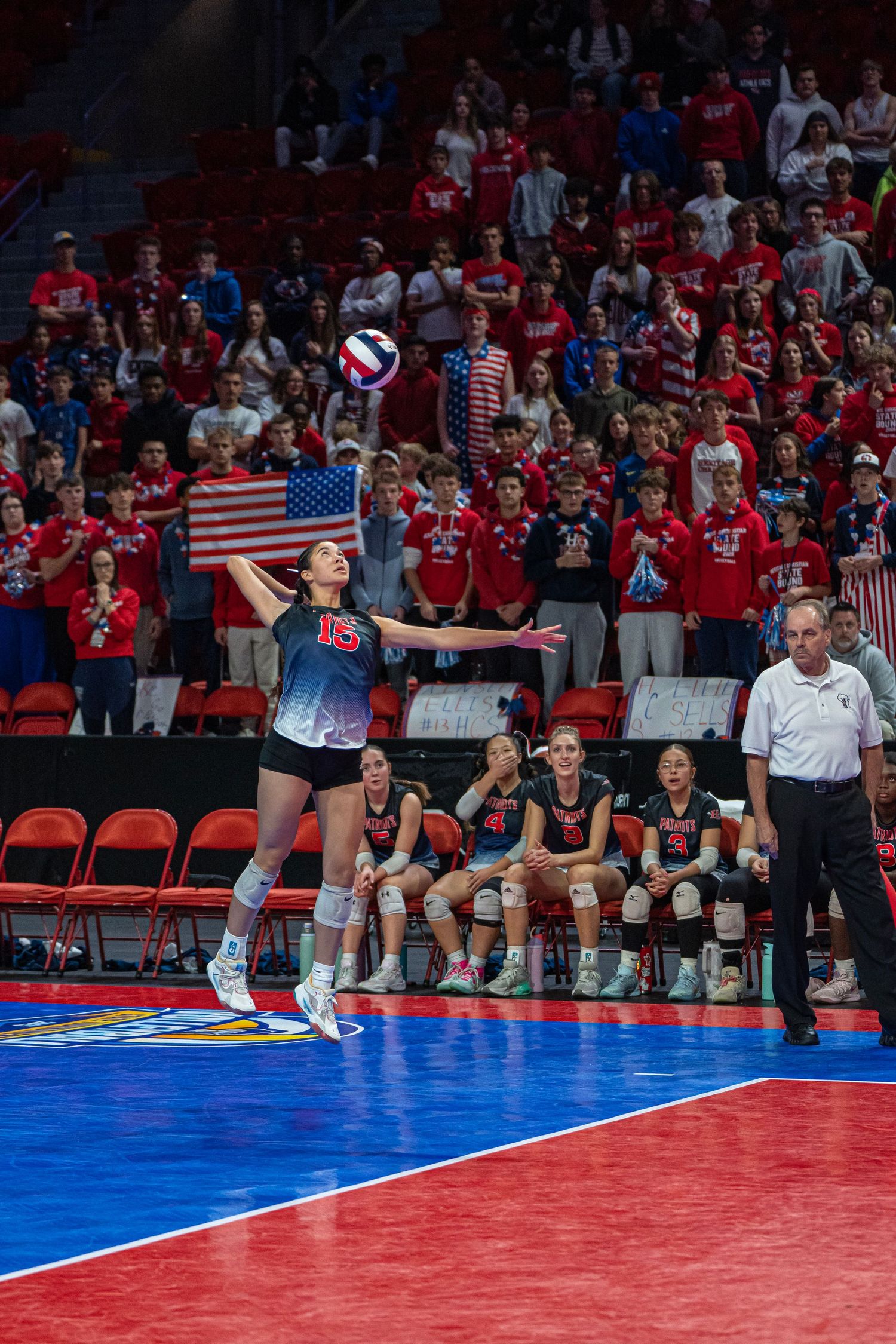 Volleyball player serves during match with red and blue court and American flag-waving crowd in background.