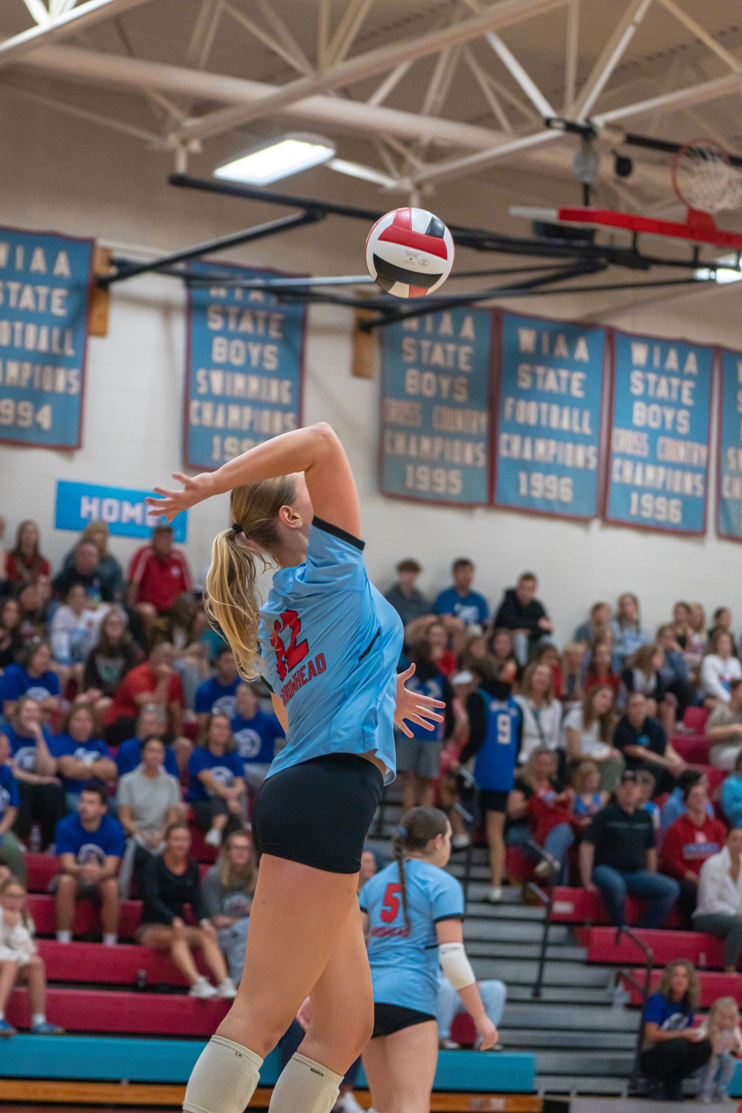 High school volleyball players compete at the net with championship banners hanging in the background.