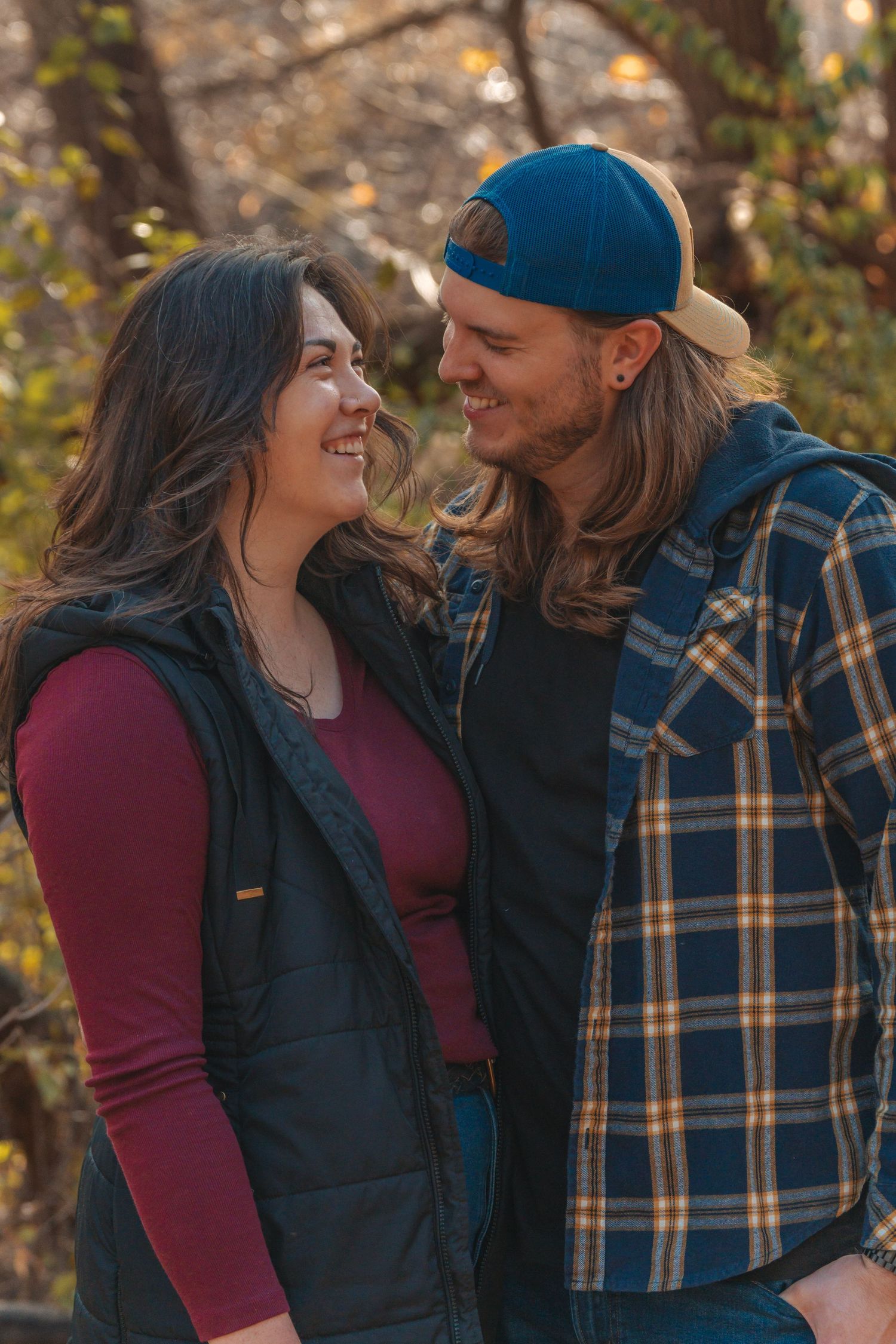 A smiling couple gazes at each other outdoors in autumn, wearing casual flannel and vest layers.