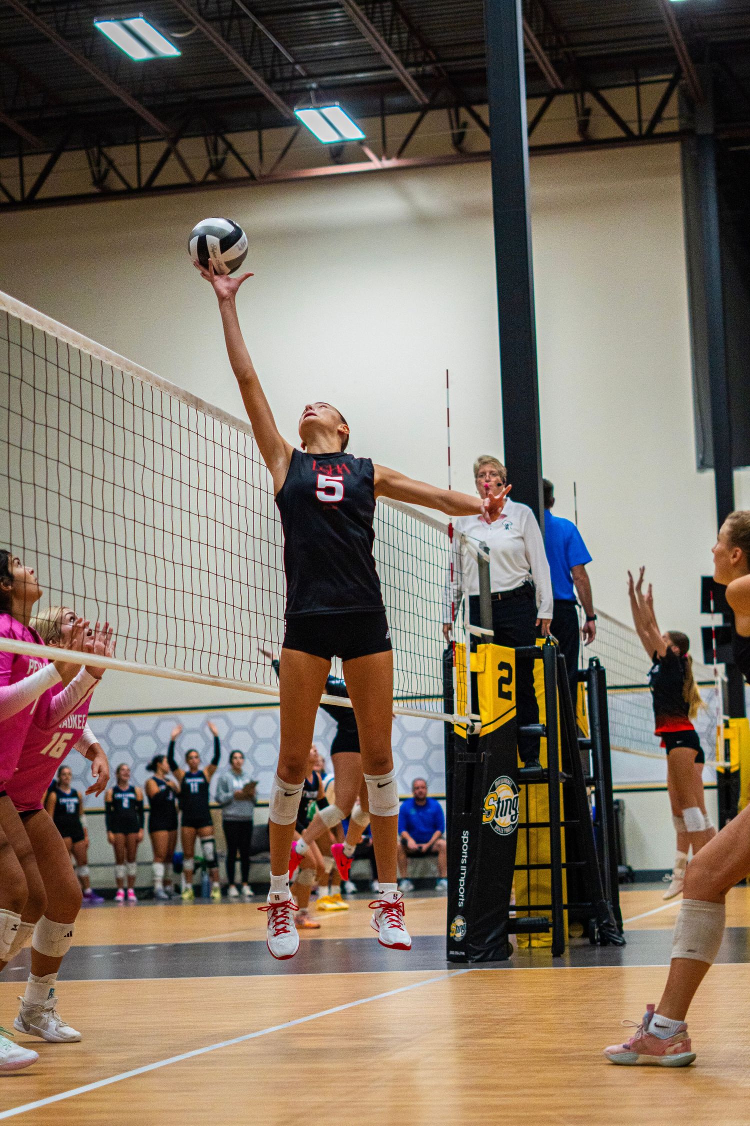 Indoor volleyball match showing players at net preparing to block and spike.