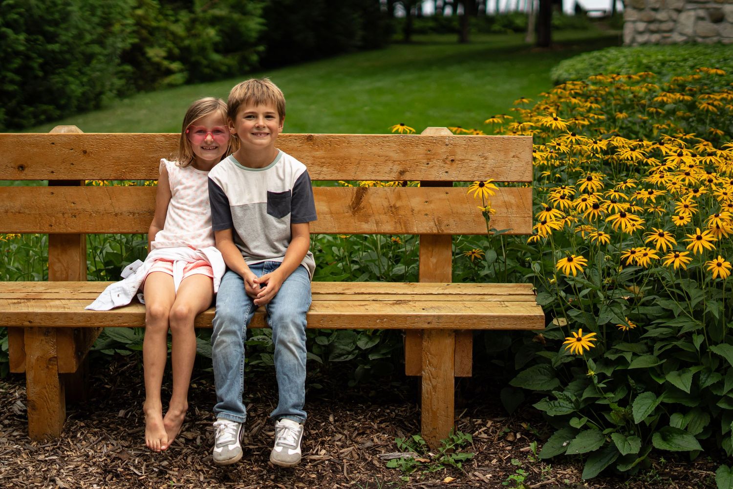 Children sitting on wooden benches surrounded by blooming yellow flowers in a garden setting.