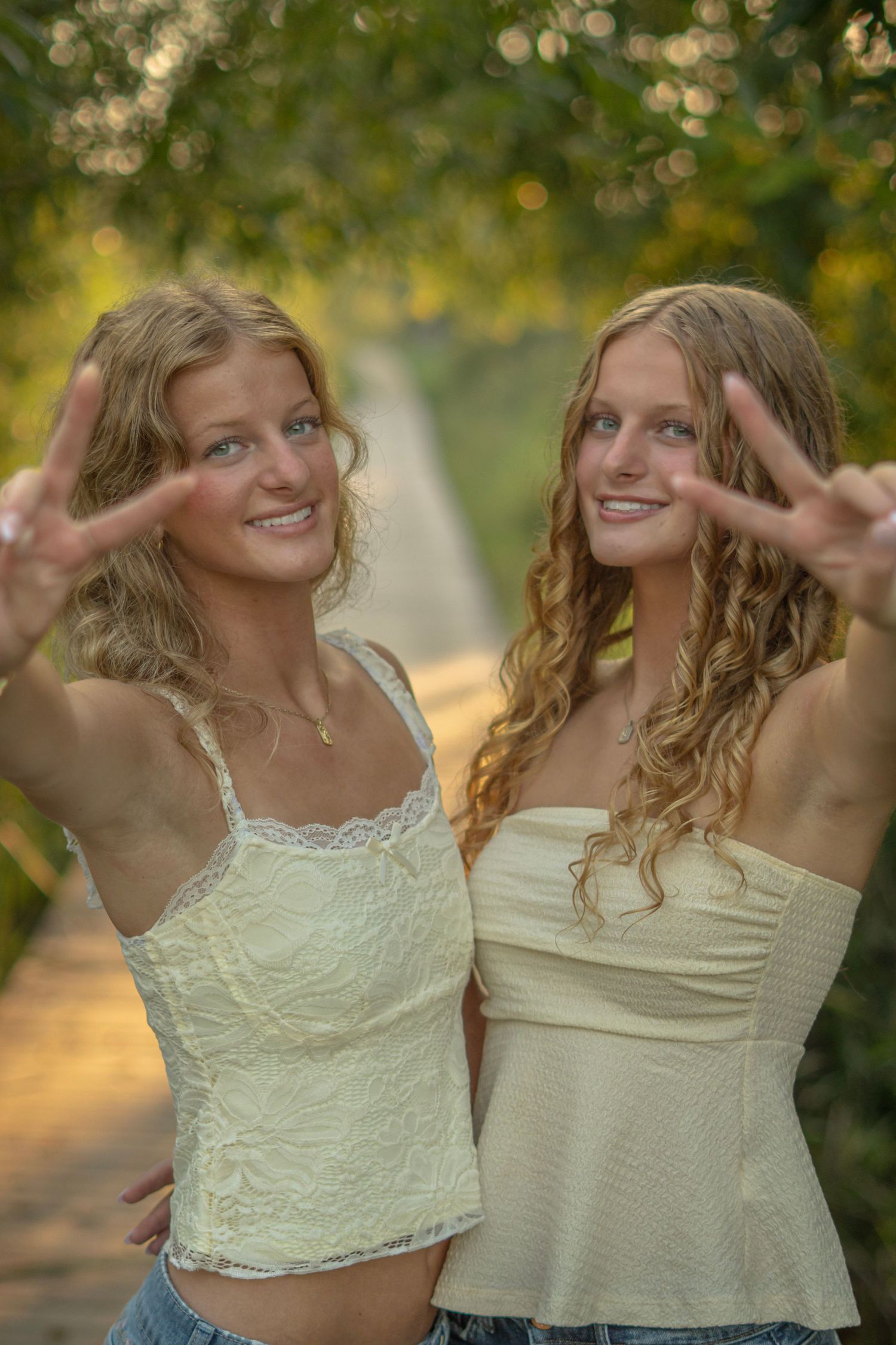Two friends in light colored tops pose playfully together outdoors with natural bokeh lighting in the background.