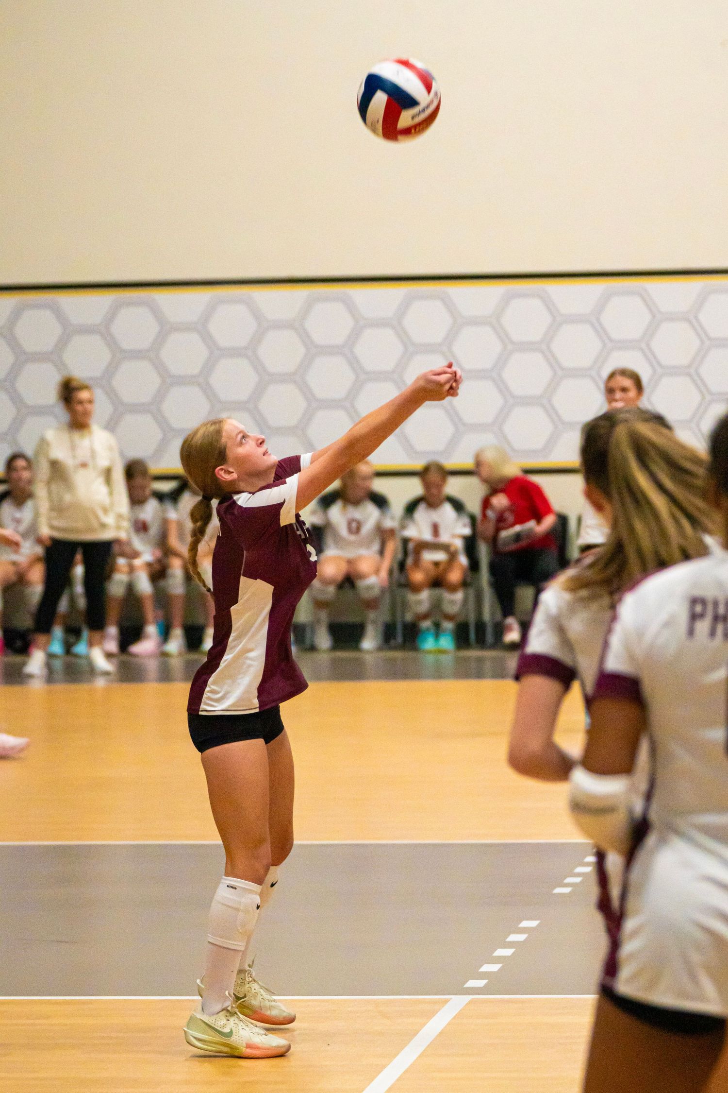 Volleyball player in maroon uniform serves ball during match.