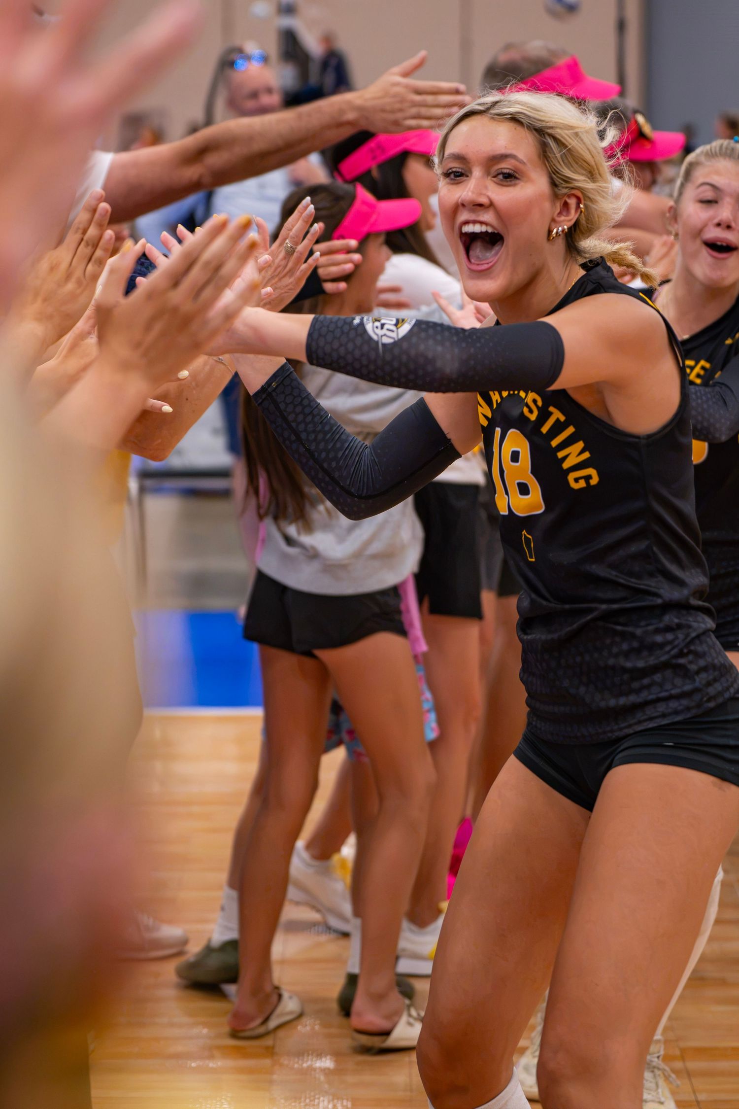Volleyball player #18 high-fives fans while entering the court, wearing a black jersey and arm sleeves.