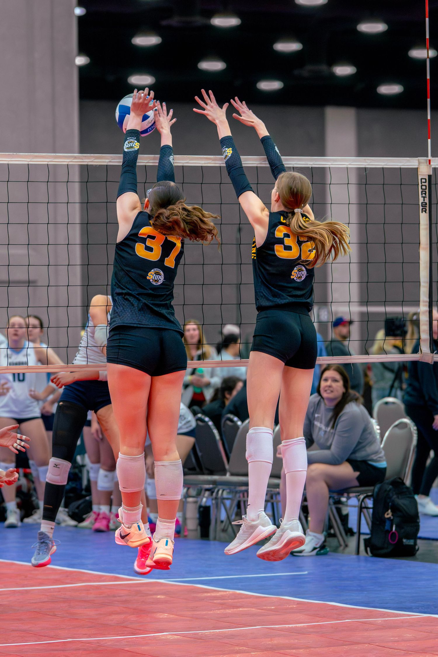 Two volleyball players in black and gold uniforms jump to block at the net during an indoor tournament match.