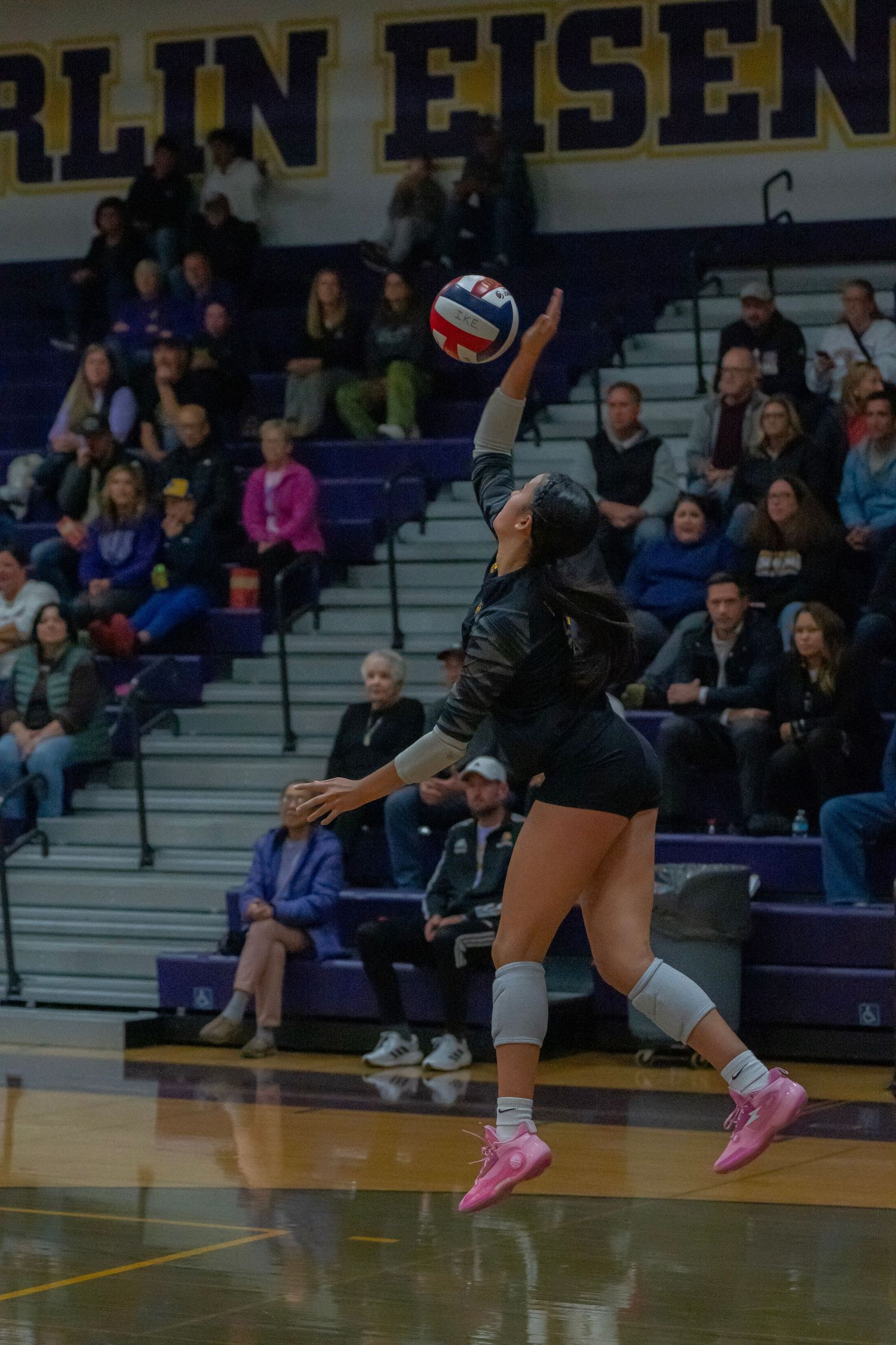 Volleyball player in black uniform stretches low to save a ball during an indoor match with spectators in background.