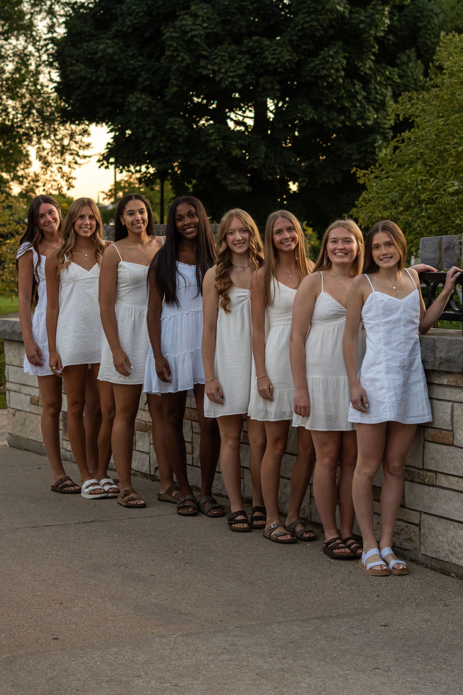 Line of people wearing matching white outfits standing against a stone wall at dusk.