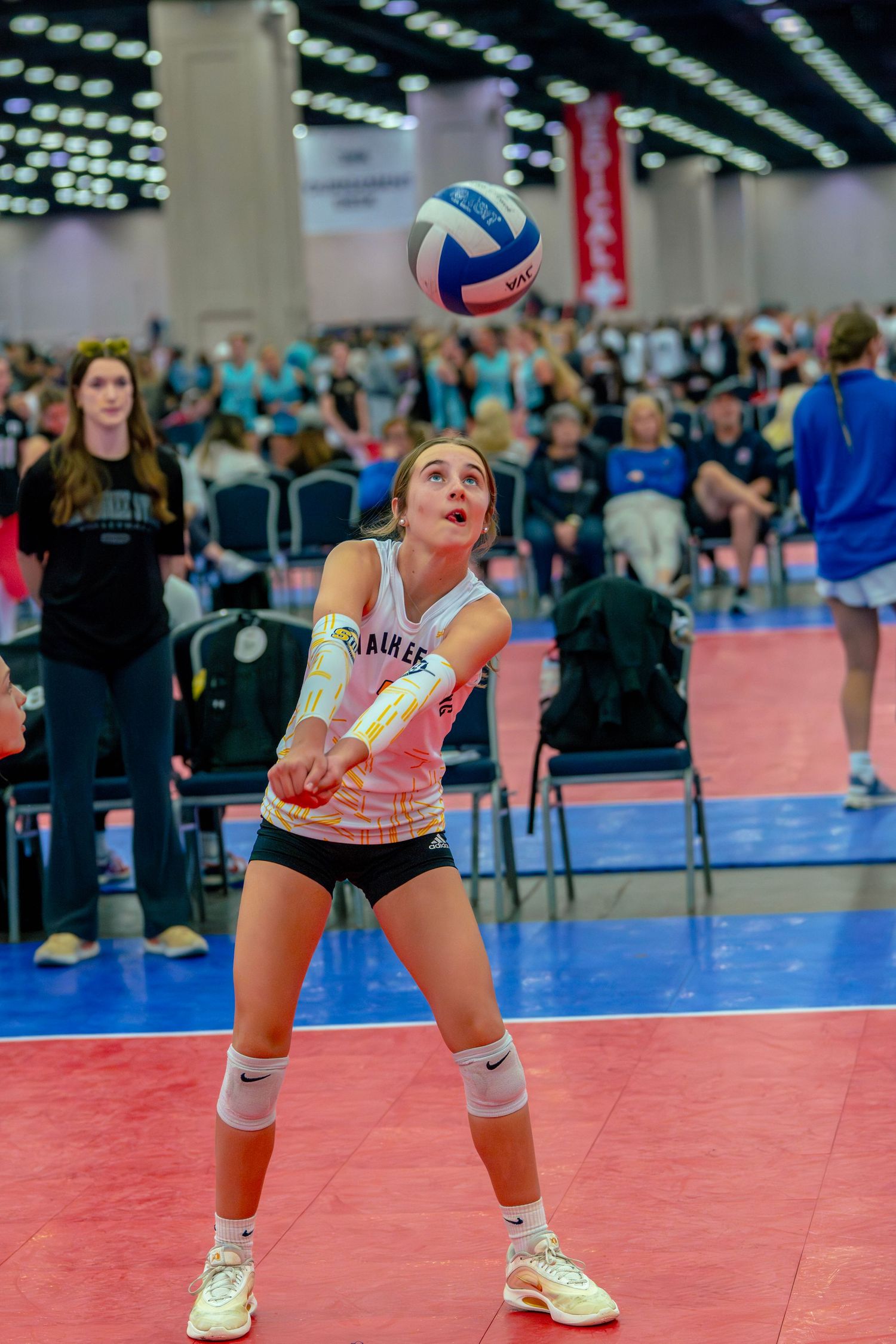 Young female volleyball player in Nike uniform bumping ball at indoor tournament with crowded arena in background.