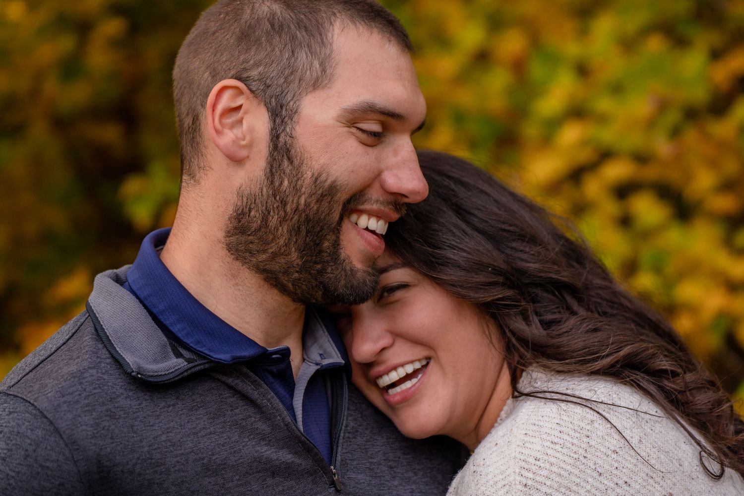 Happy couple laughing together outdoors in autumn, man with beard nuzzling smiling brunette woman among fall foliage.