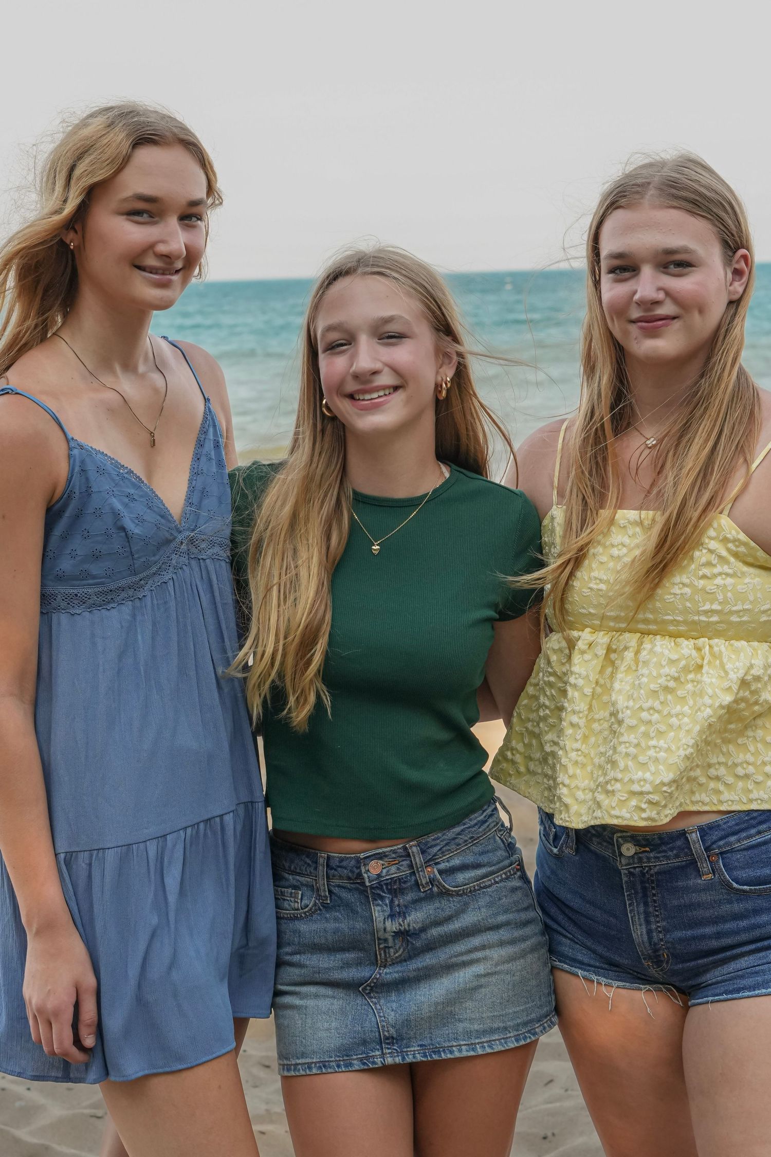 Sisters in summer outfits posing together at beach with ocean in background.