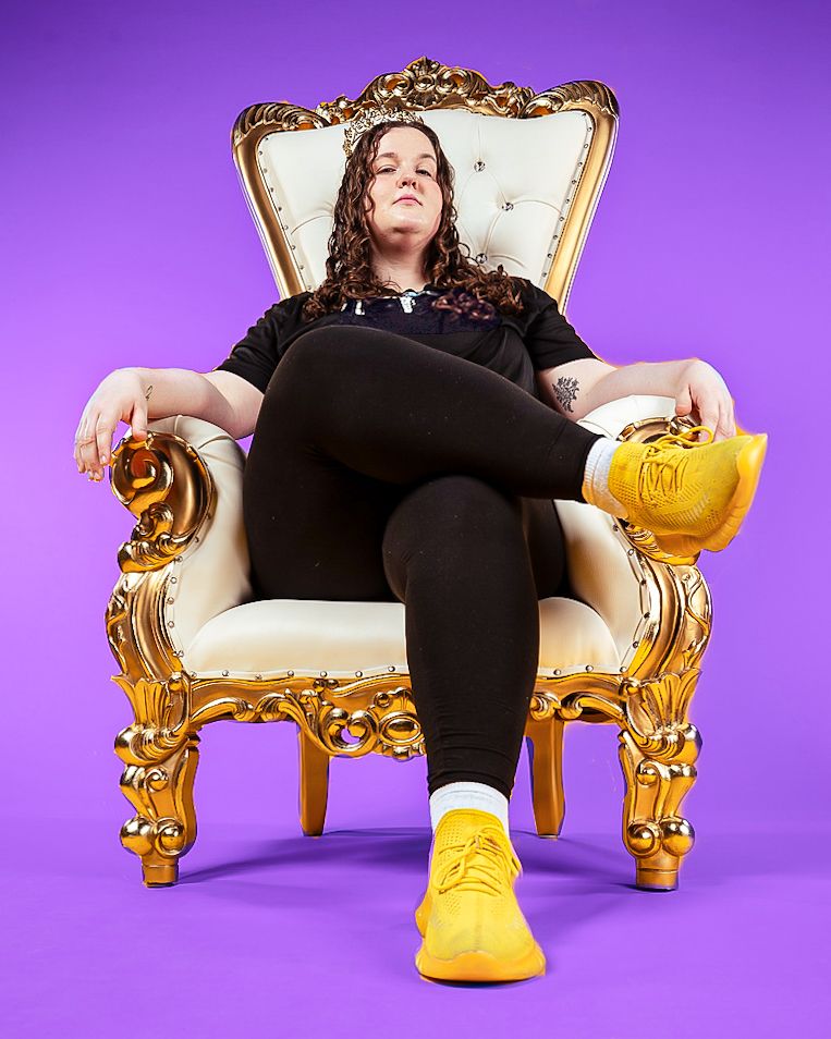 Person lounging in ornate gold chair wearing black outfit and yellow boots against purple background.