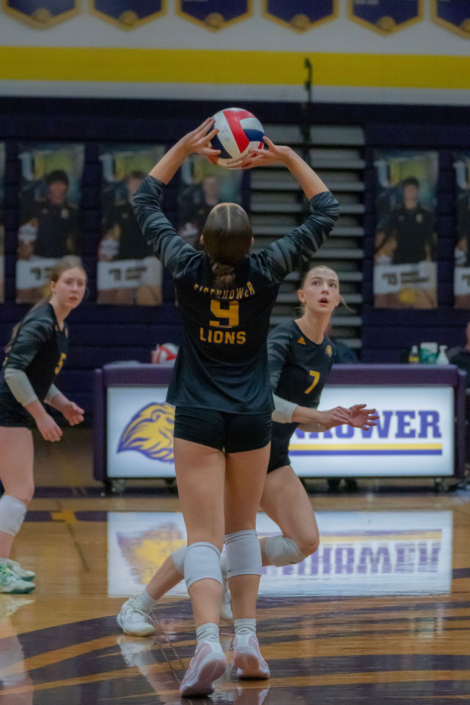 Volleyball players in black uniforms diving and reaching for the ball during an intense match on a court.