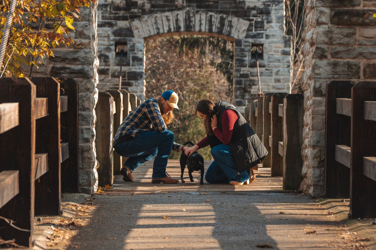 Two people crouching to pet a small black dog on a rustic stone bridge pathway in autumn.