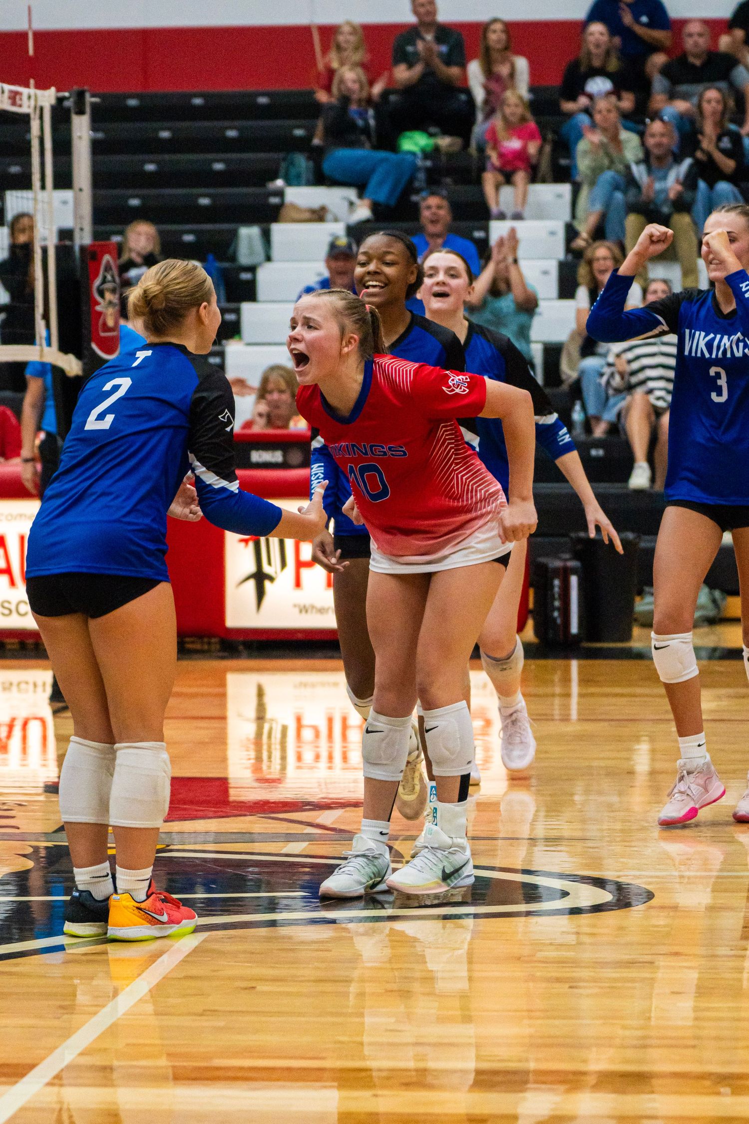 Volleyball players in red and blue uniforms kneel on a shiny gymnasium court during a competitive match.