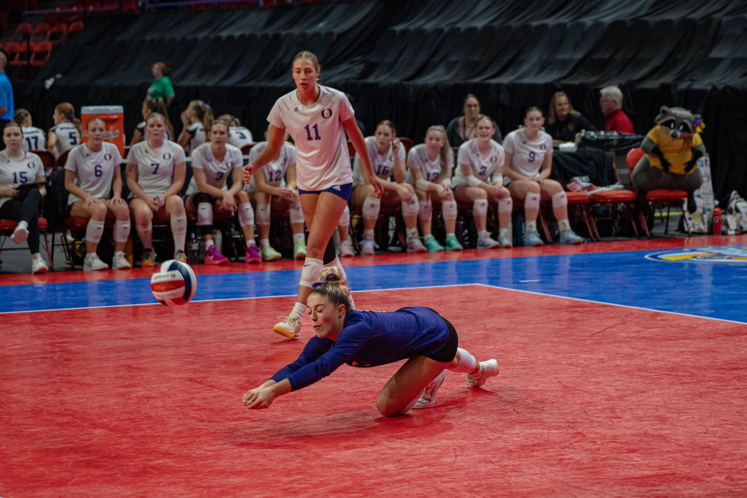 A volleyball player dives to save the ball during an intense match on a red court.