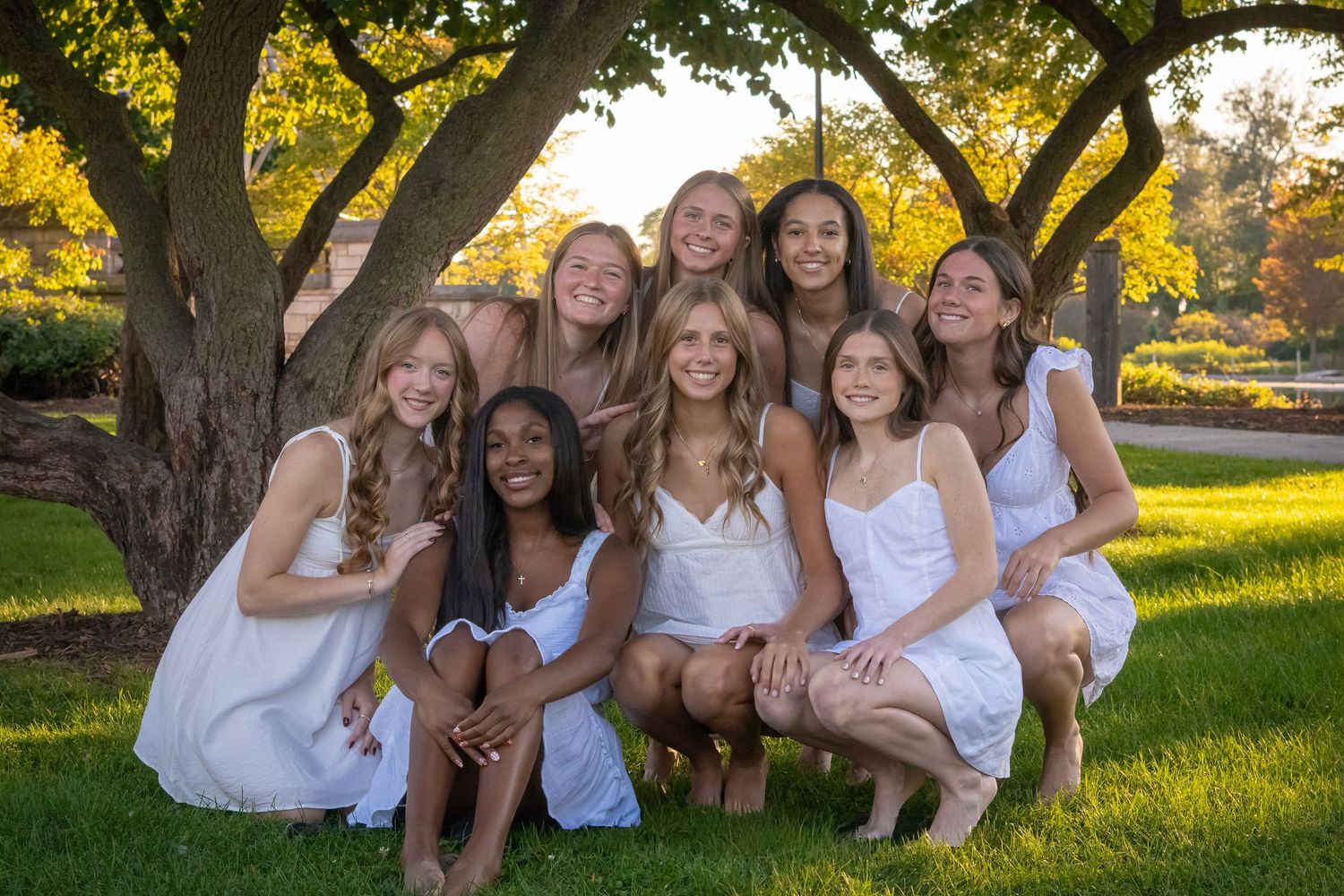 Group photo of people in white outfits sitting under a large tree in golden sunlight.