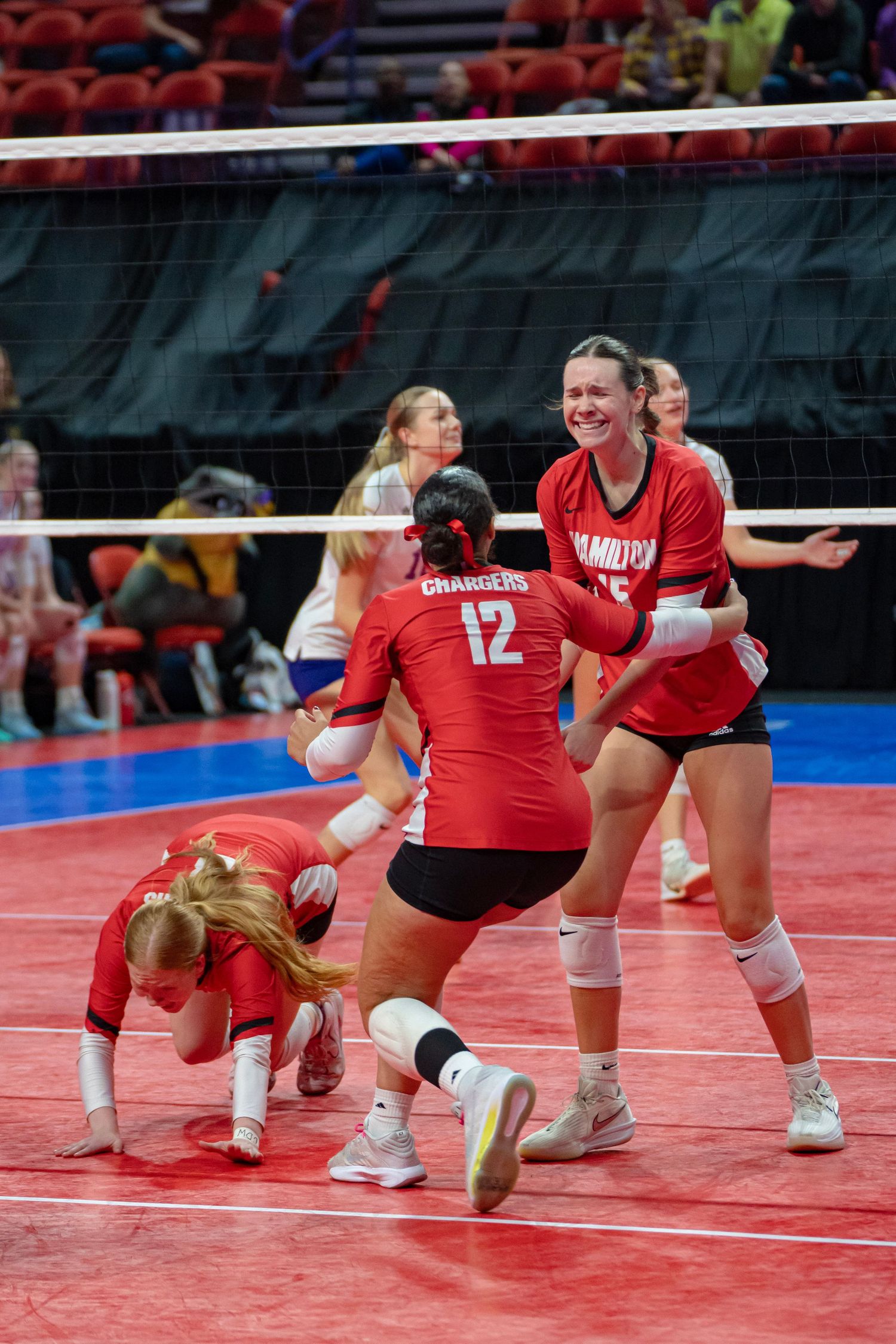 Volleyball players in red uniforms dive and stretch to save a ball during an intense match on a red court.