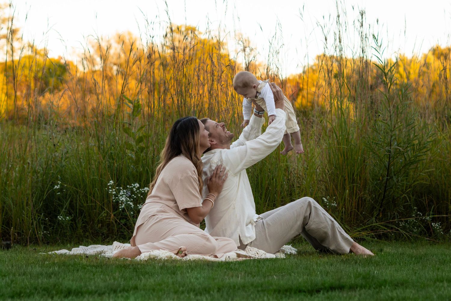A couple in white clothing sits together in a grassy meadow at sunset during a romantic photoshoot.
