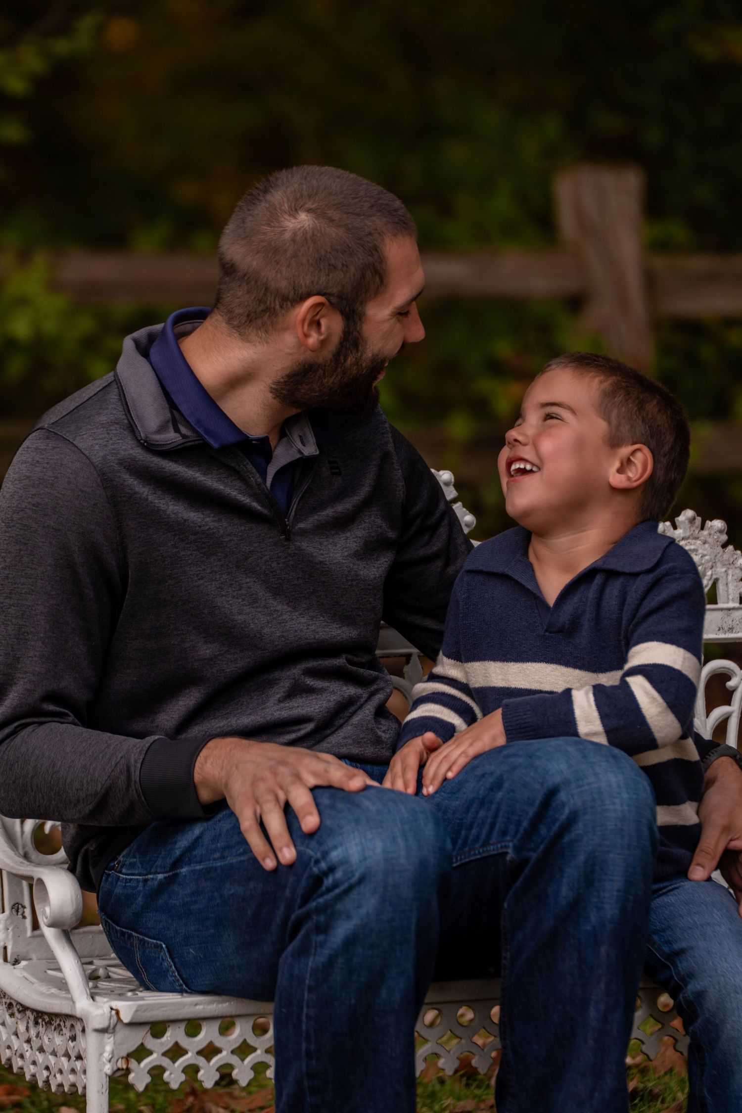 A bearded man and young boy share a joyful laugh while sitting together on a white bench outdoors.
