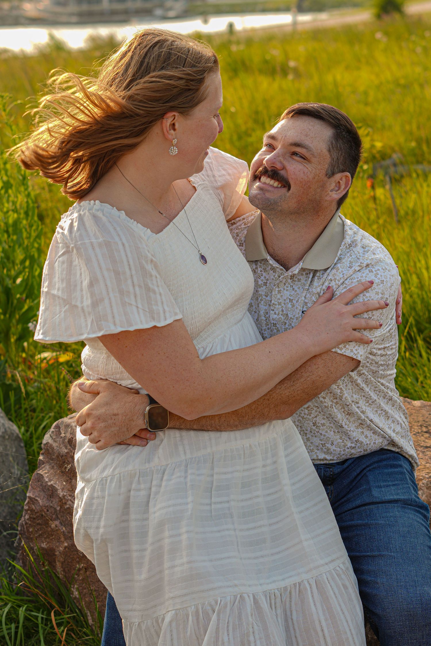 A romantic couple embraces in a sunlit meadow wearing white and casual attire.