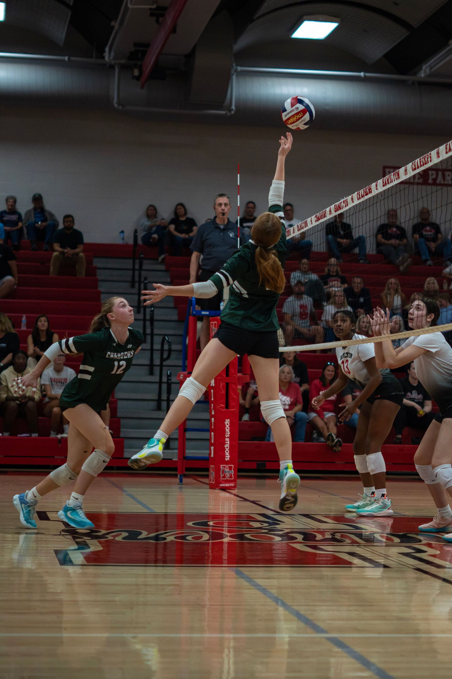 Indoor volleyball players diving and reaching for the ball during an intense game.