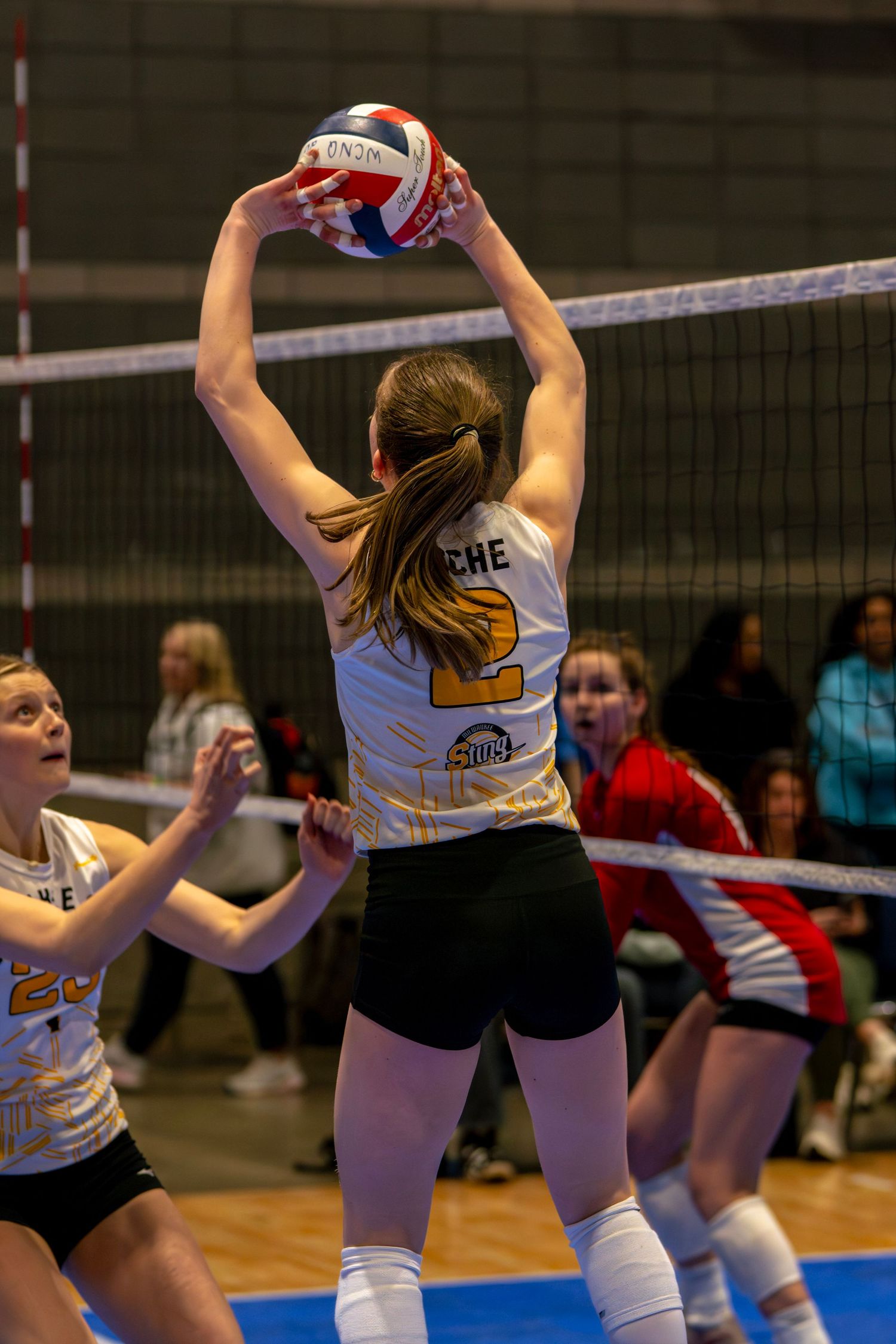 Female volleyball player #2 sets the ball during an indoor match, with teammates and opponents visible near the net.