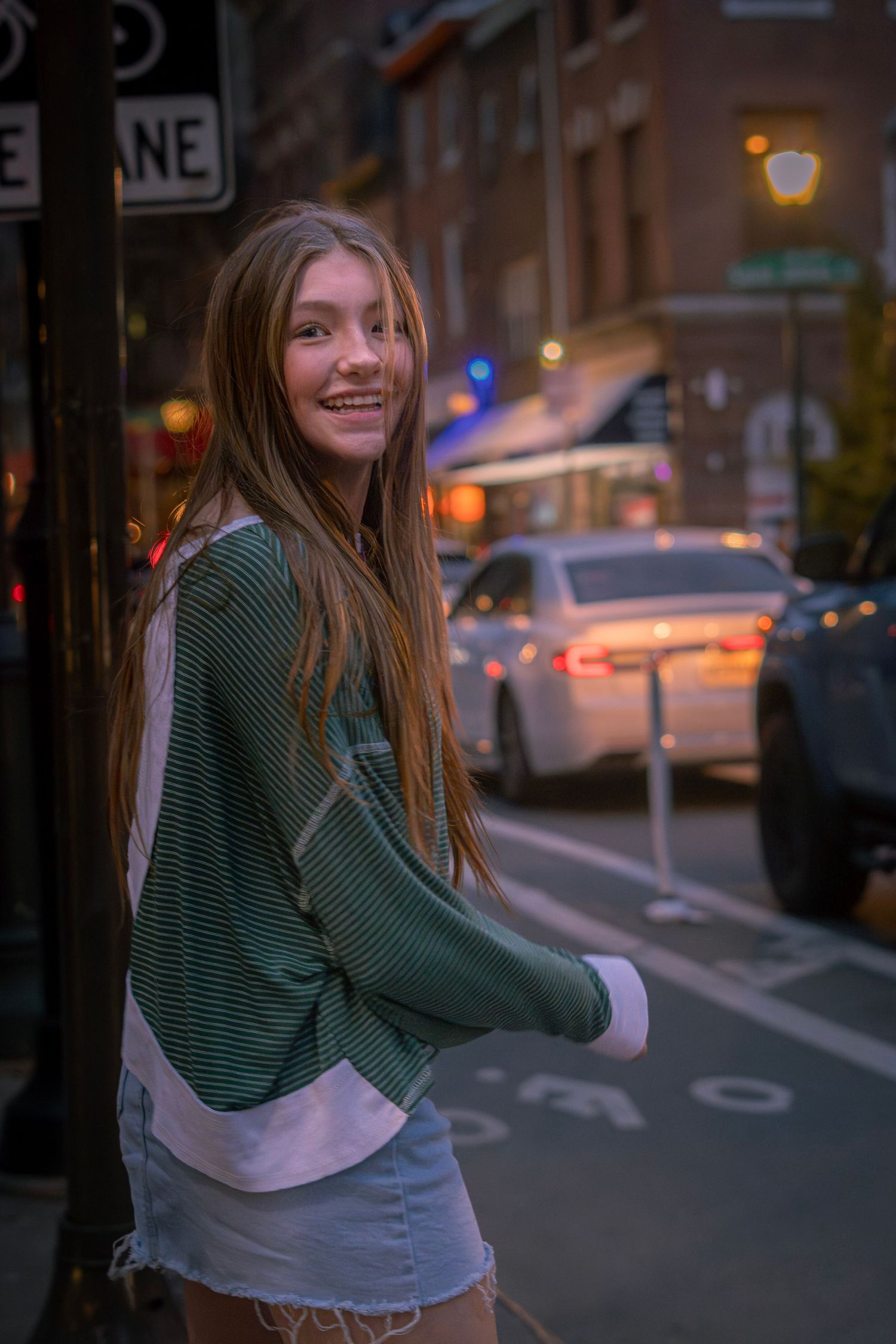 Smiling young woman in green striped top and denim shorts posing on a city street at dusk with cars and lights behind her.