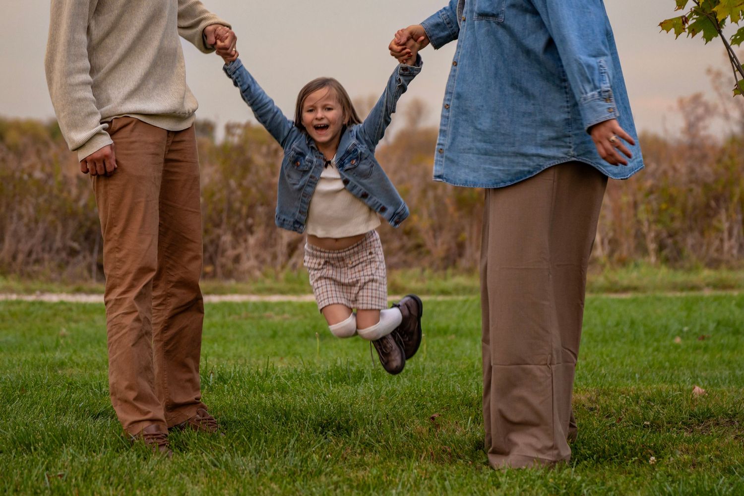 A person holds and swings a small child by their hands in a grassy outdoor setting during autumn.