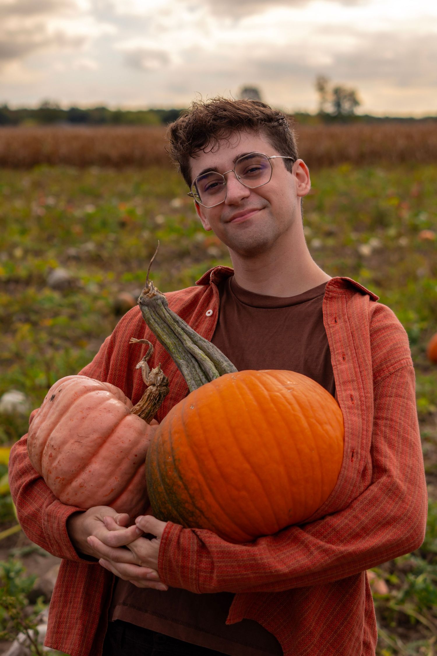 Someone wearing an orange sweater holds a large orange pumpkin in a field during autumn harvest season.