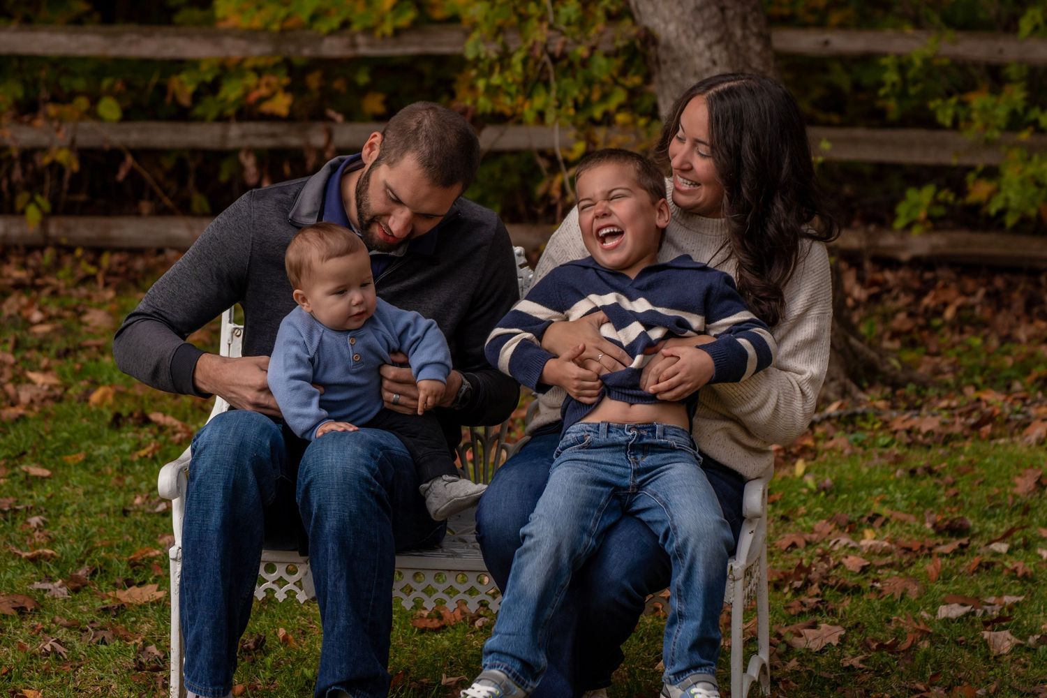 Family of four sitting on white bench outdoors in autumn, parents laughing with two young boys in matching blue outfits.