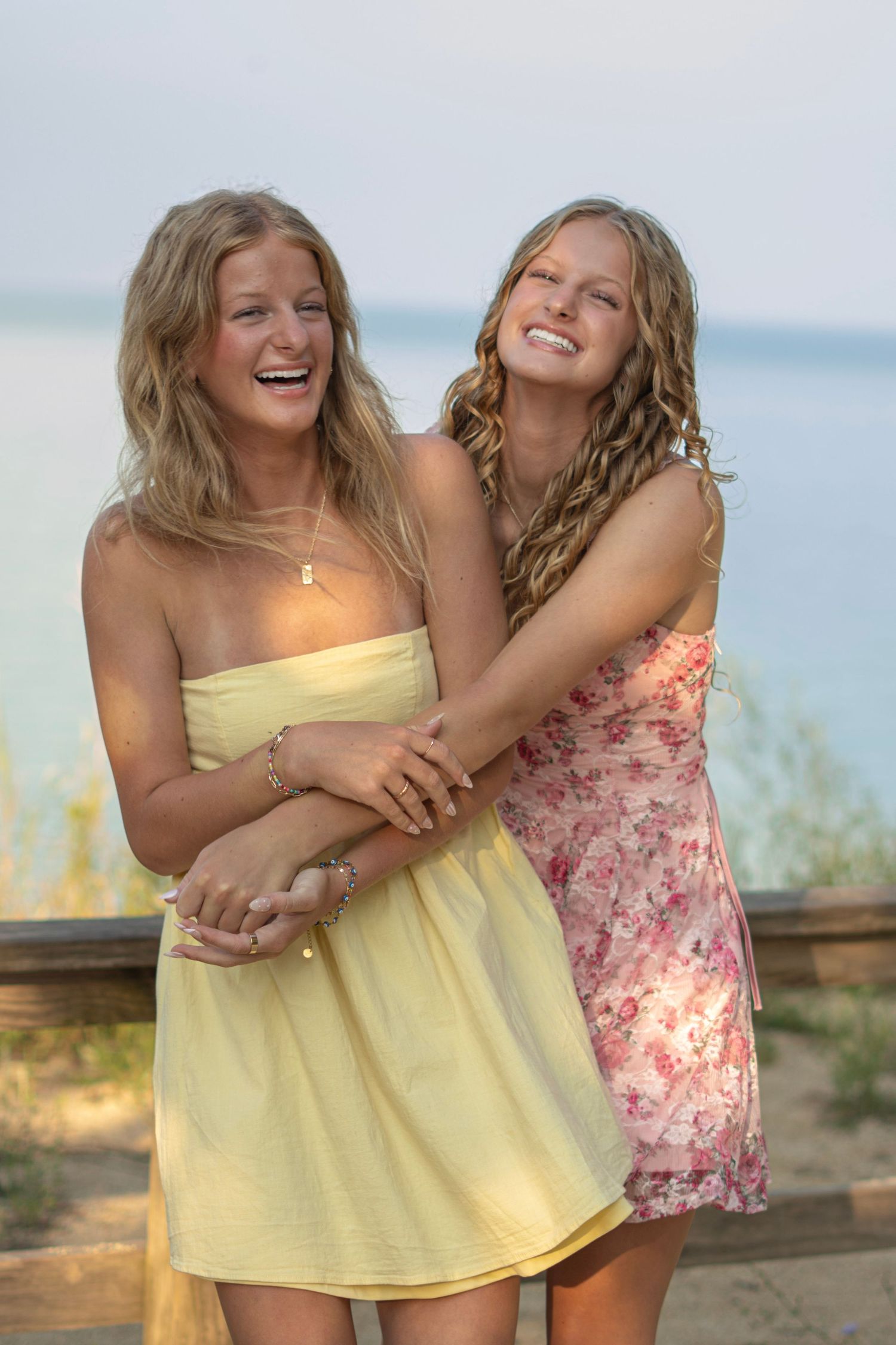 Twins embrace and laugh together on a beach at sunset wearing yellow and floral summer dresses.