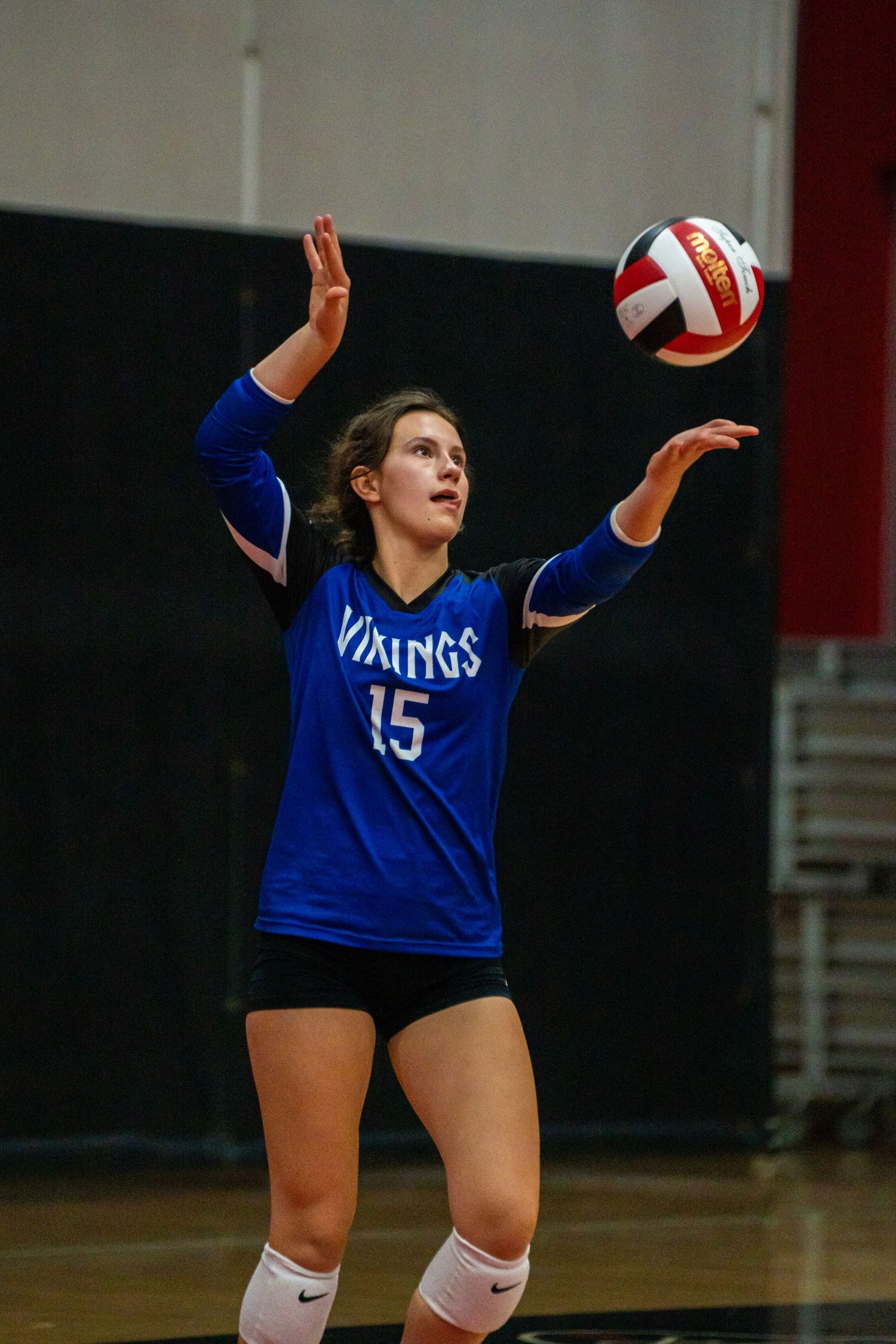 Player in blue Vikings jersey serves volleyball during competitive match.