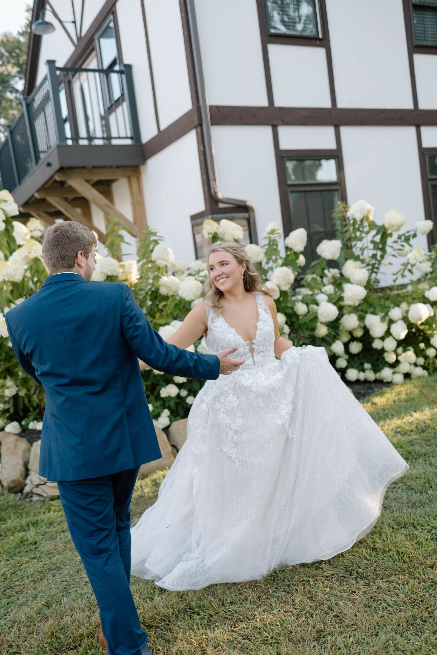 Bride and groom share playful moment on lawn in front of Tudor-style building.