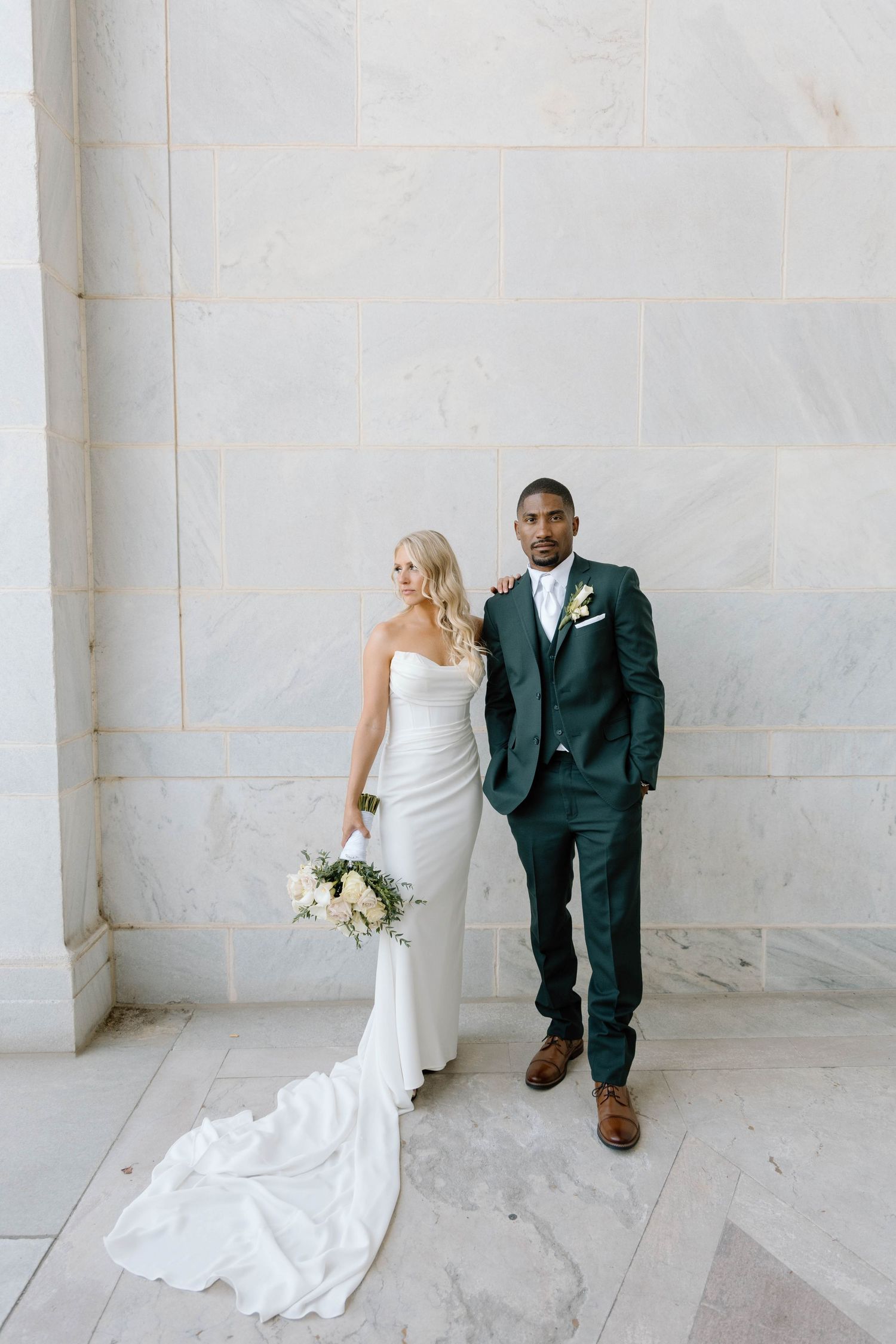 Bride and groom pose against white marble wall showcasing elegant wedding attire.