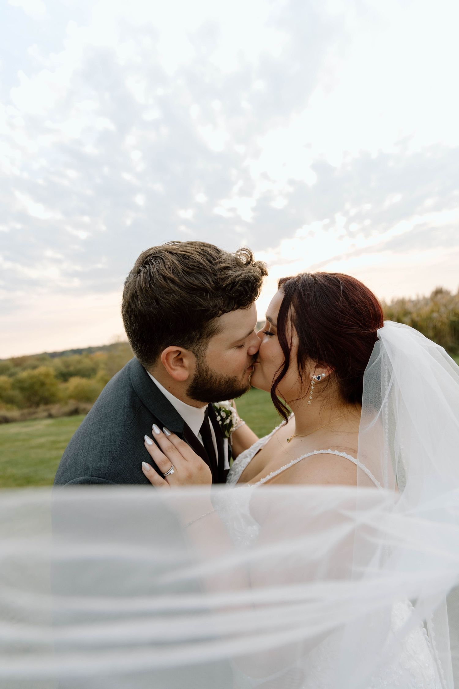 Romantic wedding couple kisses under flowing veil during sunset photo session.