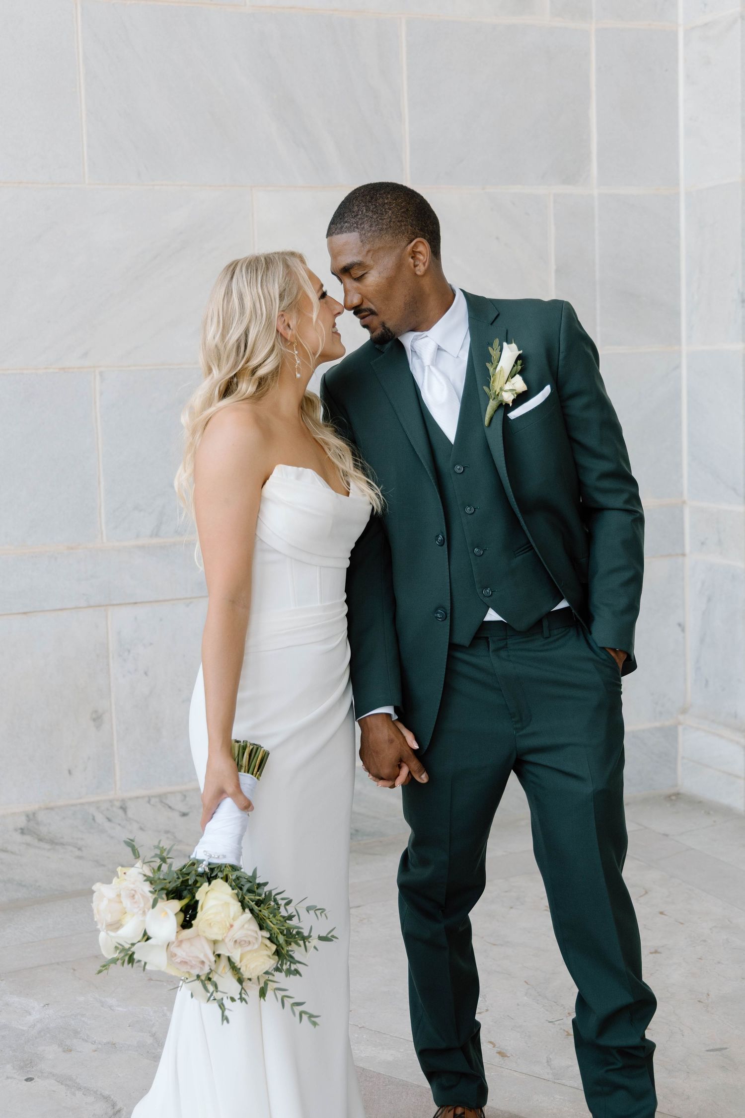 Elegant wedding couple poses against a stone wall with the bride holding a white floral bouquet.