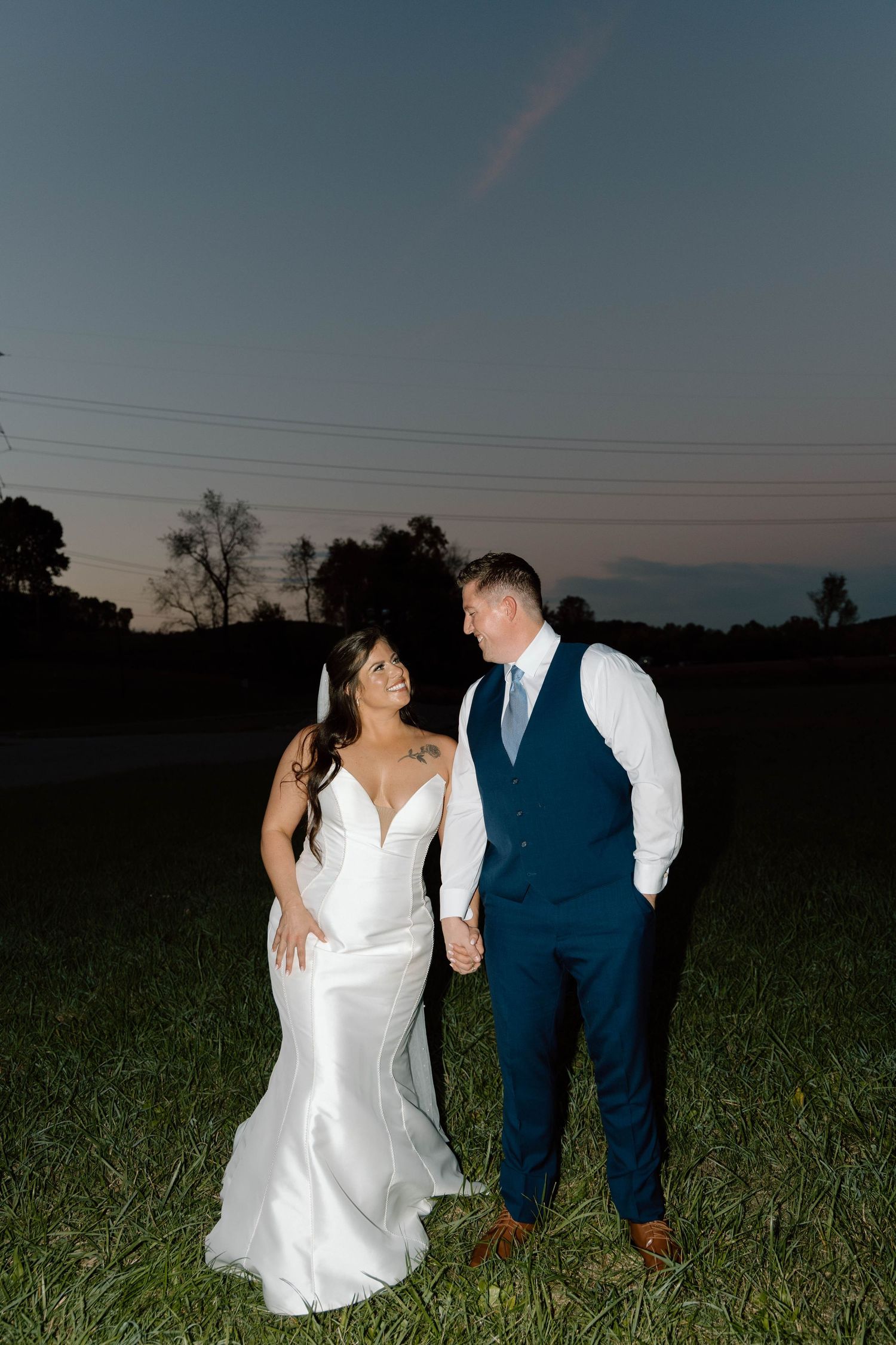 Newlyweds pose together in a grassy field at dusk wearing formal wedding attire.
