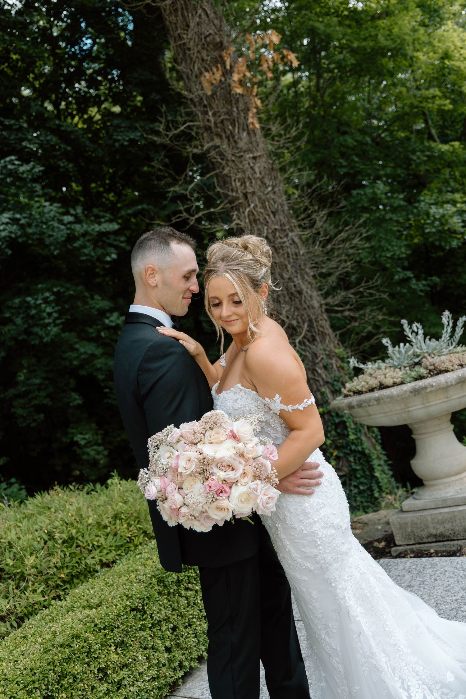 Wedding couple embraces in garden setting surrounded by lush greenery and flowers.